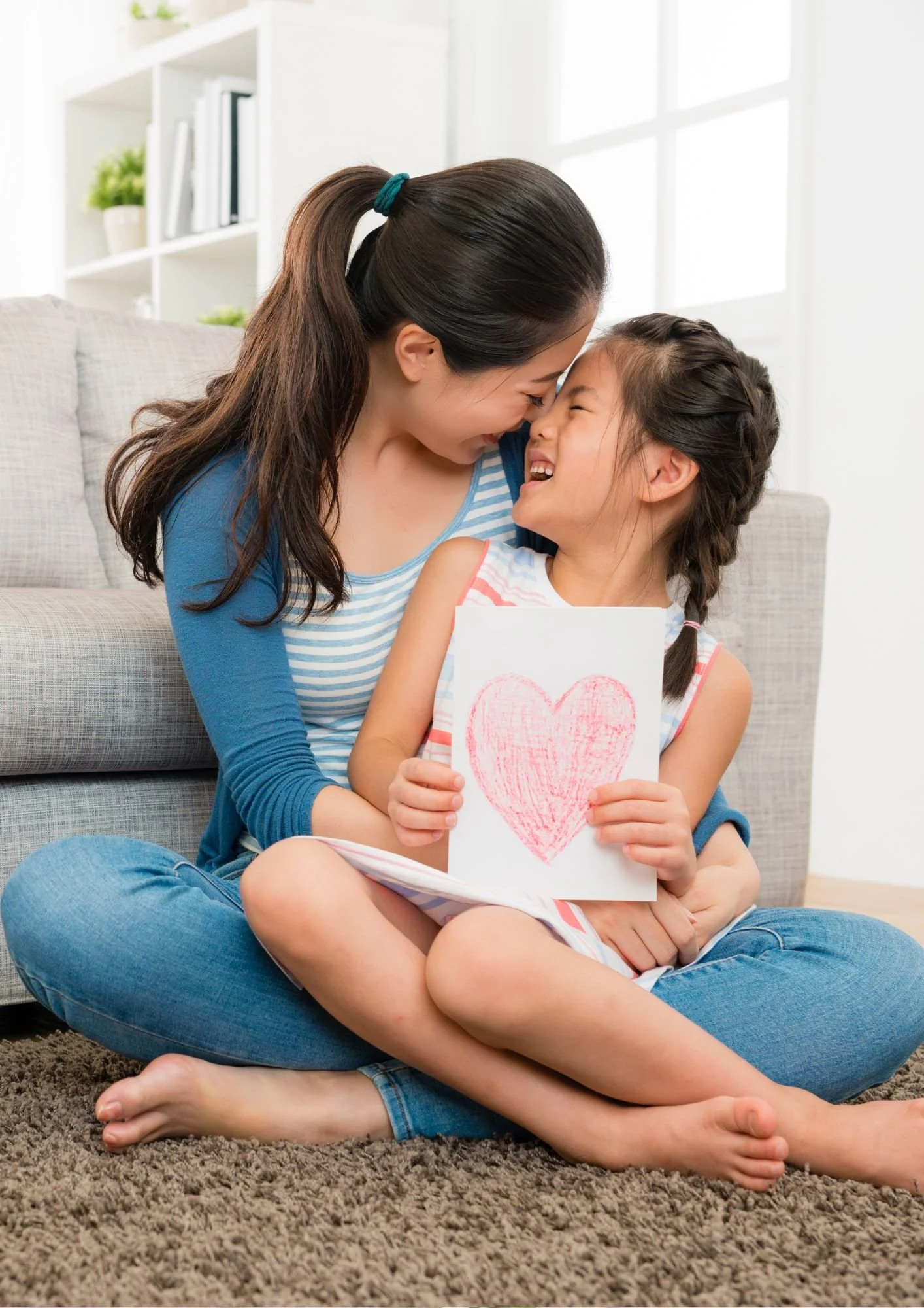 A mother and daughter sitting on a carpeted floor, smiling and touching foreheads. The daughter is holding a drawing of a red heart.