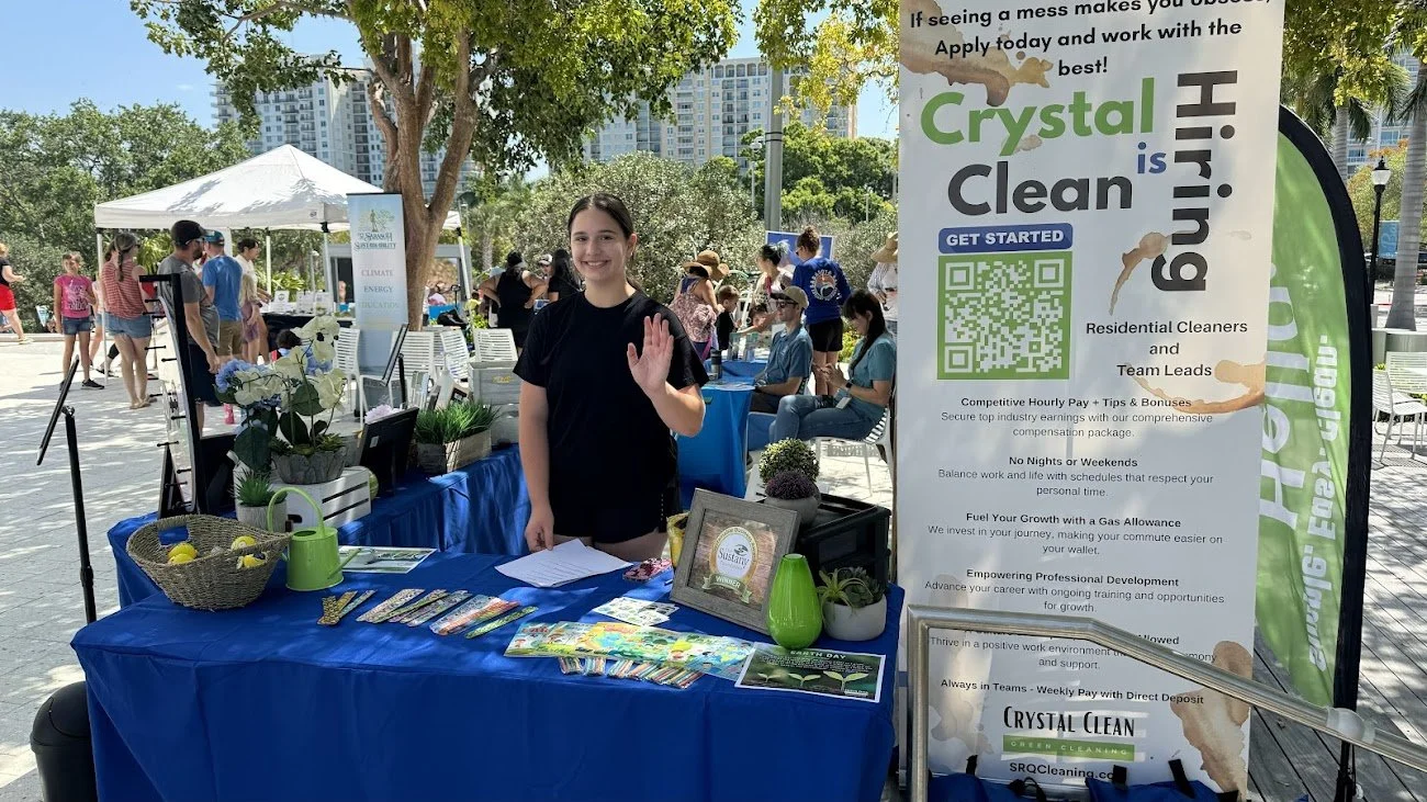A young woman is standing at a table with a blue tablecloth at an outdoor event, smiling and waving. The table has promotional materials, a small potted plant, and a green watering can. Behind her, other people are gathered around various booths under tents, with trees and tall buildings in the background. A large vertical banner promoting 'Crystal Clean' residential cleaning services is next to her.
