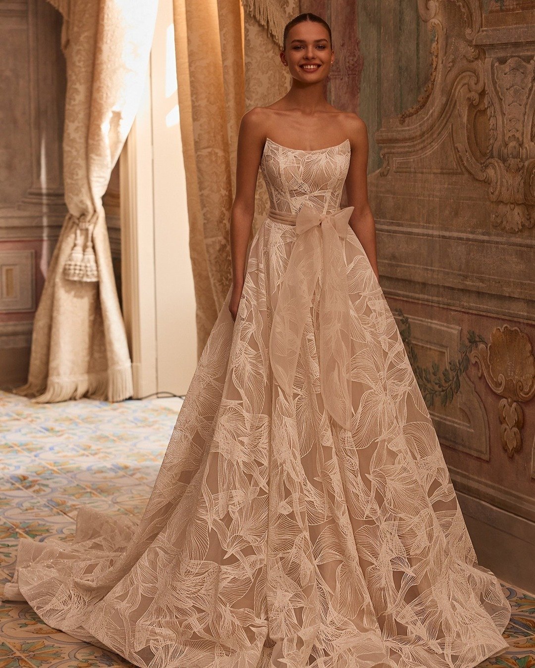 Shimmering lace, a sculpted neckline, and the dreamiest A-line silhouette ✨ This strapless gown stuns with intricate patterned lace, soft glitter details, and a flattering detachable belt to define the waist. Finished with a chapel train and delicate