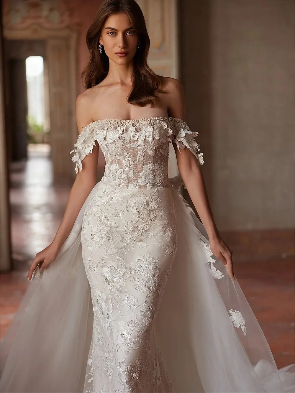 A vision of romance.
This off-the-shoulder gown stuns with 3D floral appliqu&eacute;, a sheer corset bodice, and a detachable overskirt for two unforgettable looks in one. Perfect for the bride who wants elegance with a touch of modern allure.

#bgsb