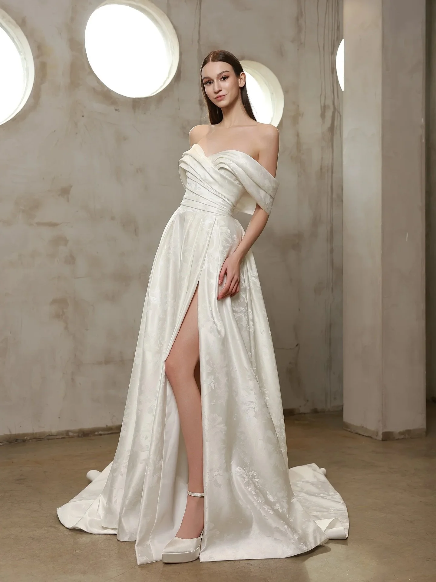 A new arrival with understated drama. This off-the-shoulder gown features a softly draped neckline, a flattering fitted waist, and a flowing A-line skirt designed to move with ease. Subtle textured fabric and a thigh-high slit bring a modern edge, wh