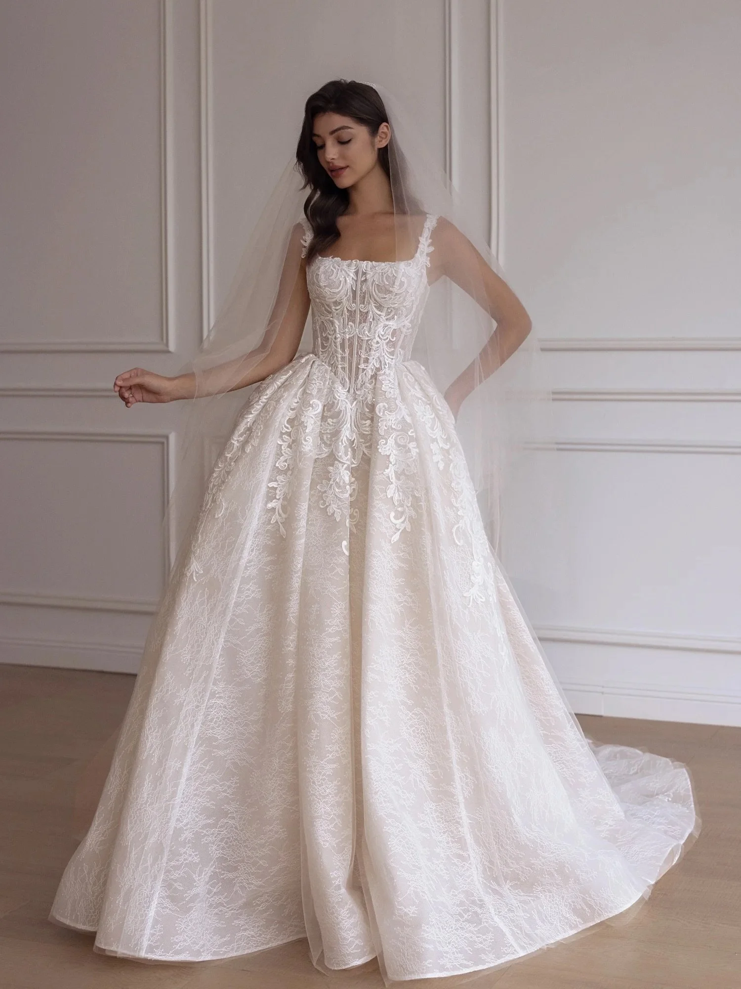 This square-neck gown features hand-placed floral lace, sculpted structure, and just the right amount of skin peeking through for a look that feels timeless, romantic, and beautifully refined.