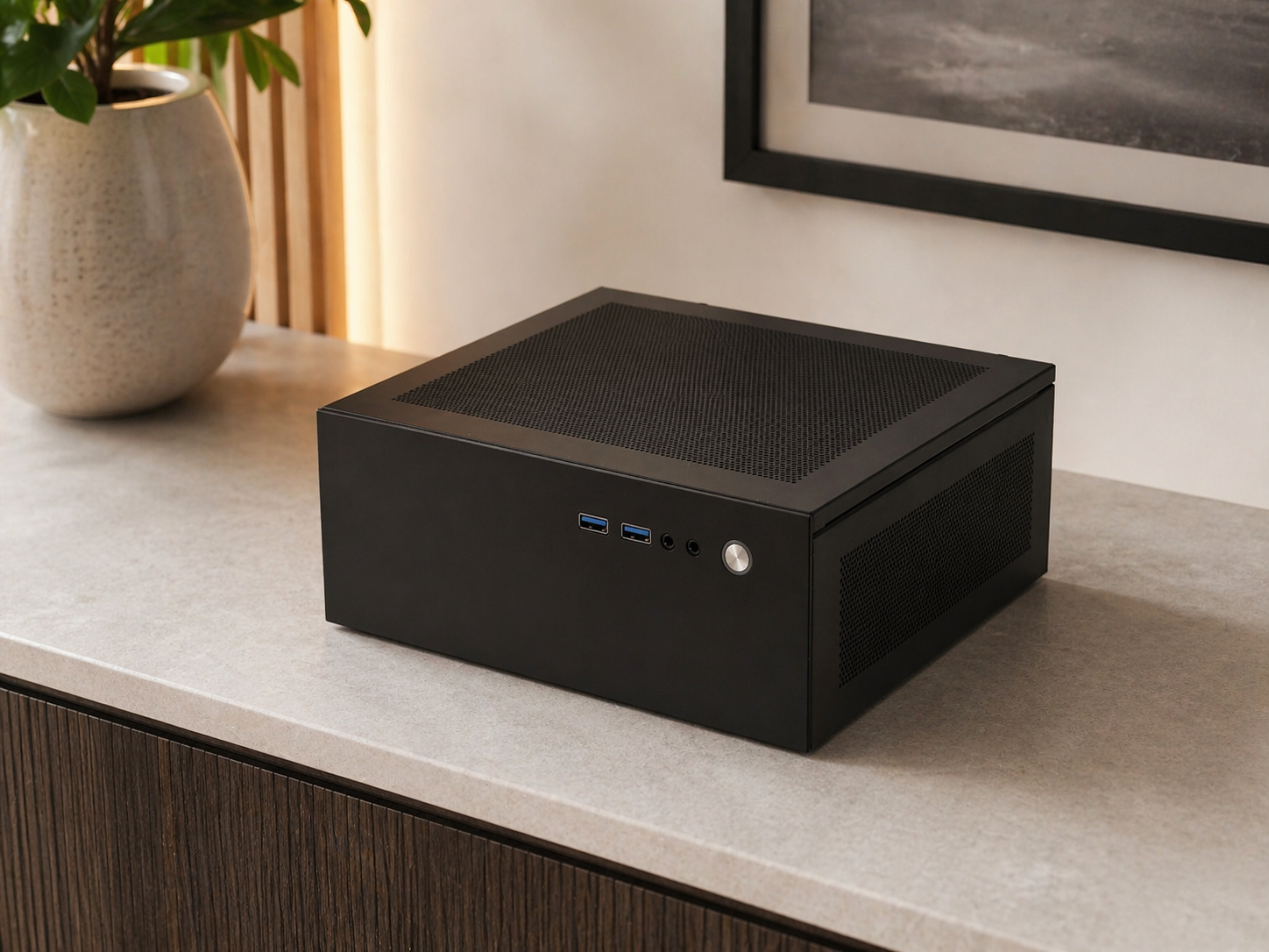A black computer case with ventilation holes, two USB ports, a headphone jack, and a power button, placed on a light-colored table near a potted plant and framed artwork.