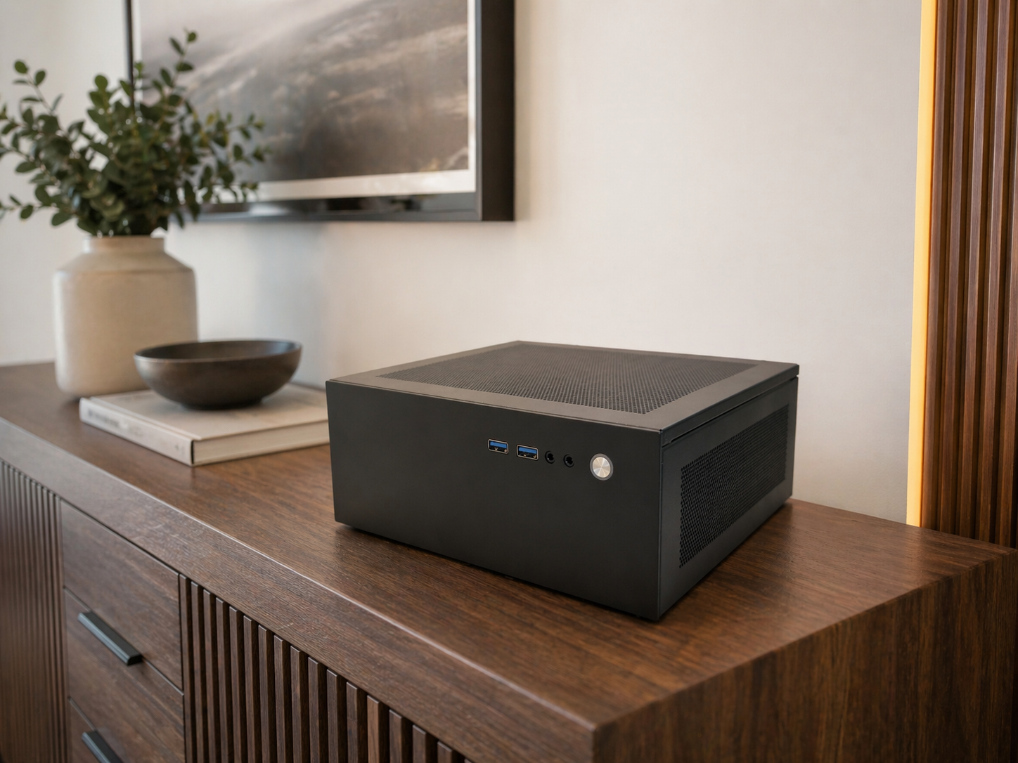 Black mini PC with front USB ports and power button on wooden sideboard, beside a potted plant, bowl, and art display on the wall.