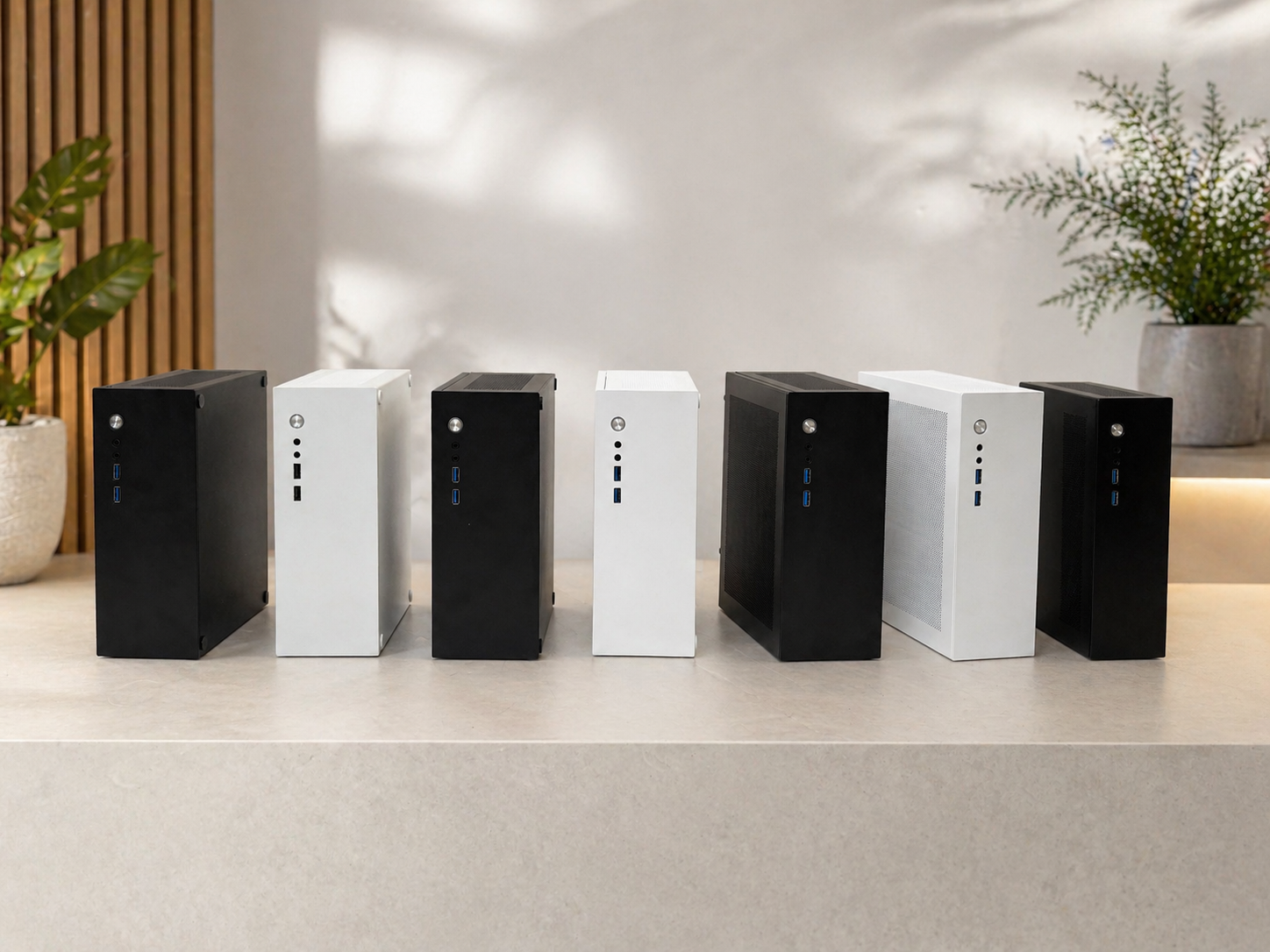 Six small desktop computers in black and white with USB ports and power buttons, lined up on a light-colored table, with potted plants and a wooden slat wall in the background.