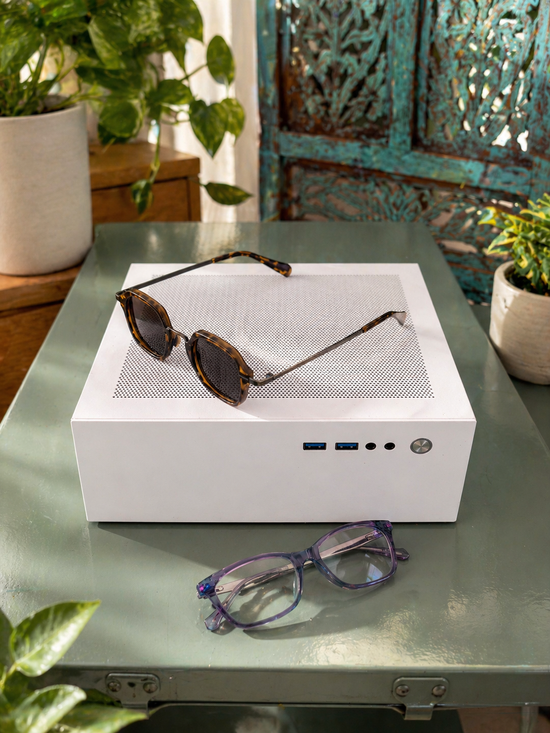 A white computer case with sunglasses on top and a pair of glasses in front, placed on a glass table with plants in the background.