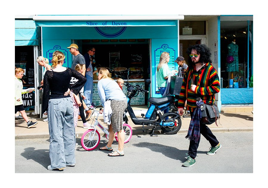 A Man Wearing Colourful Clothes and Fashion Sunglasses Passes By Families outside a Fast Food Shop. There is lots of colour in the picture