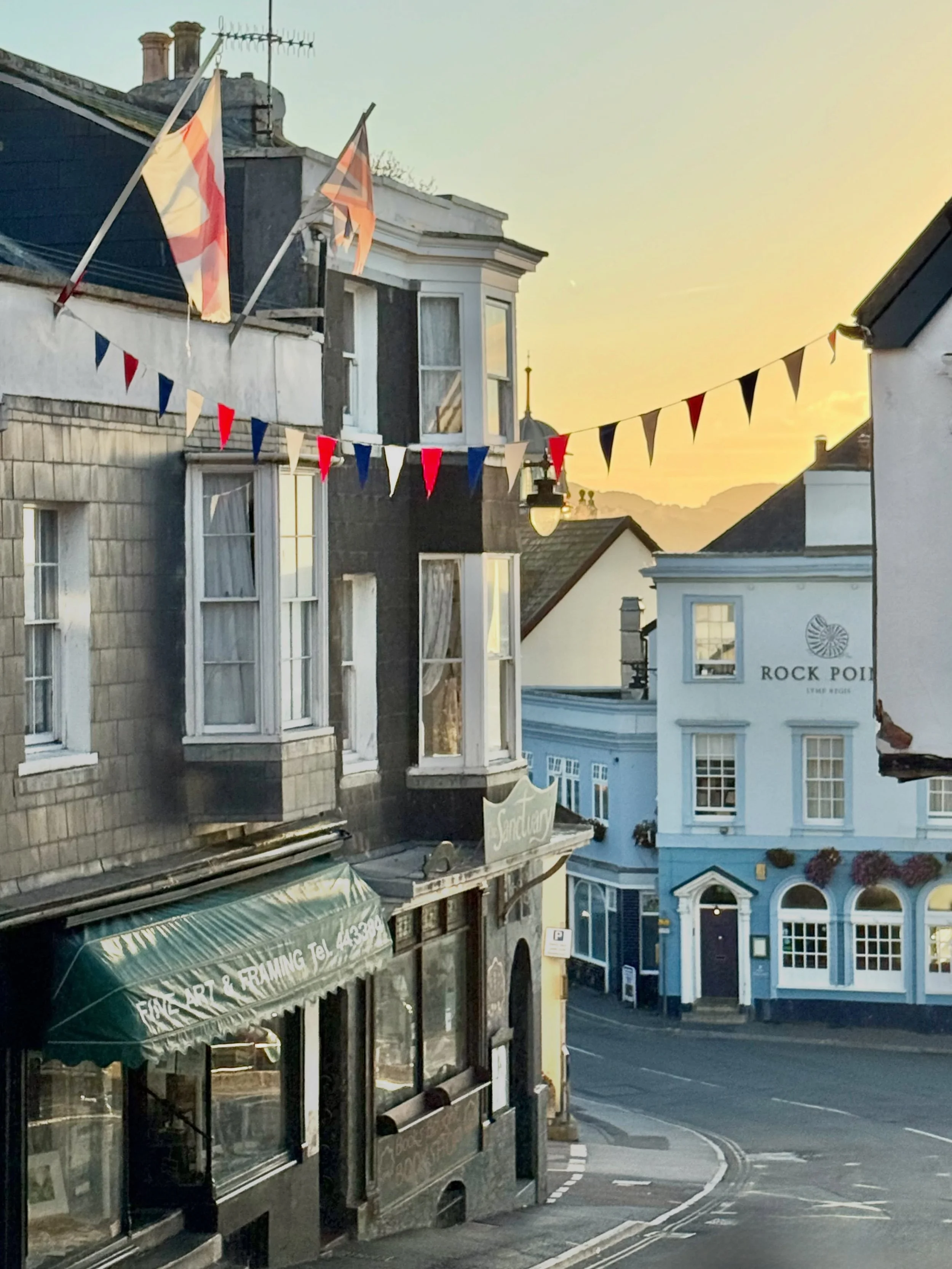 Broad Street serves as the vibrant commercial heart of Lyme Regis, sweeping steeply down from the upper town toward the seafront and offering striking views of Lyme Bay at its foot. Lined with an eclectic mix of Georgian and Victorian facades, colorf