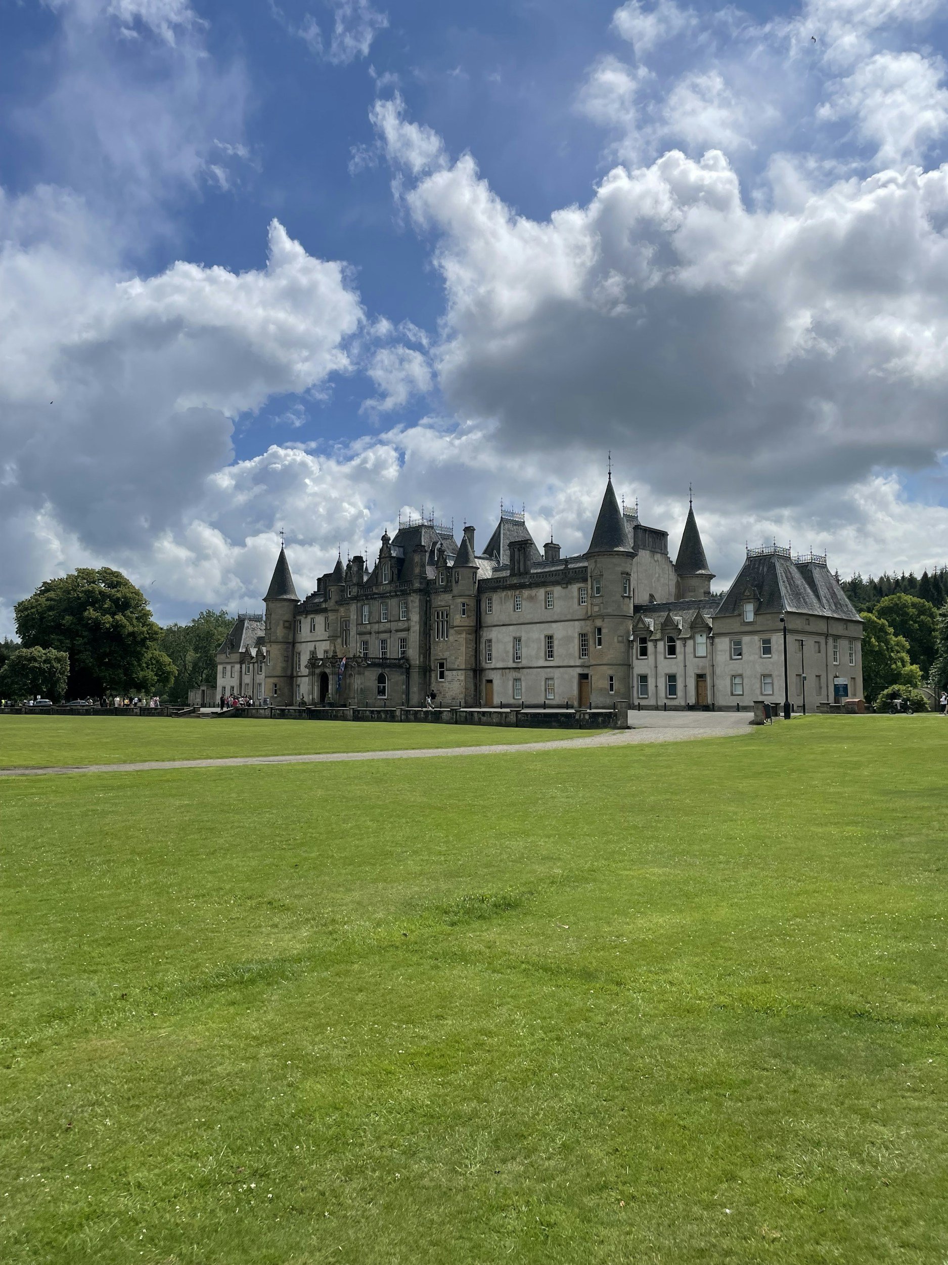 A large, historic castle with multiple turrets and towers, surrounded by a vast green lawn and a partly cloudy sky.