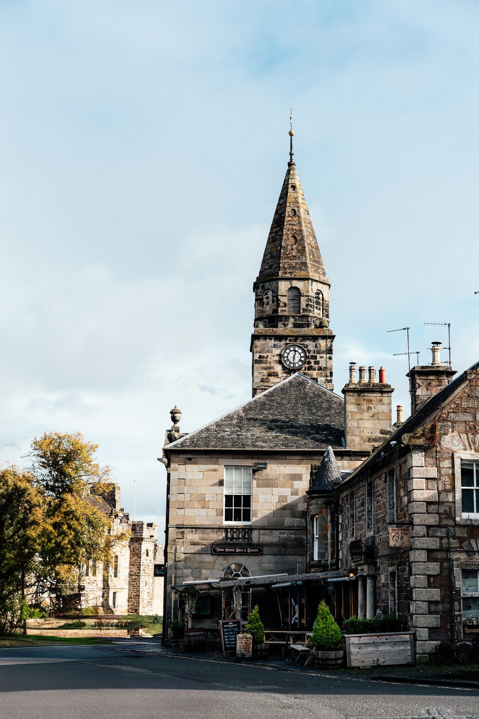 Historic stone building with a clock tower, a cafe sign, and outdoor seating, surrounded by trees and other stone structures.