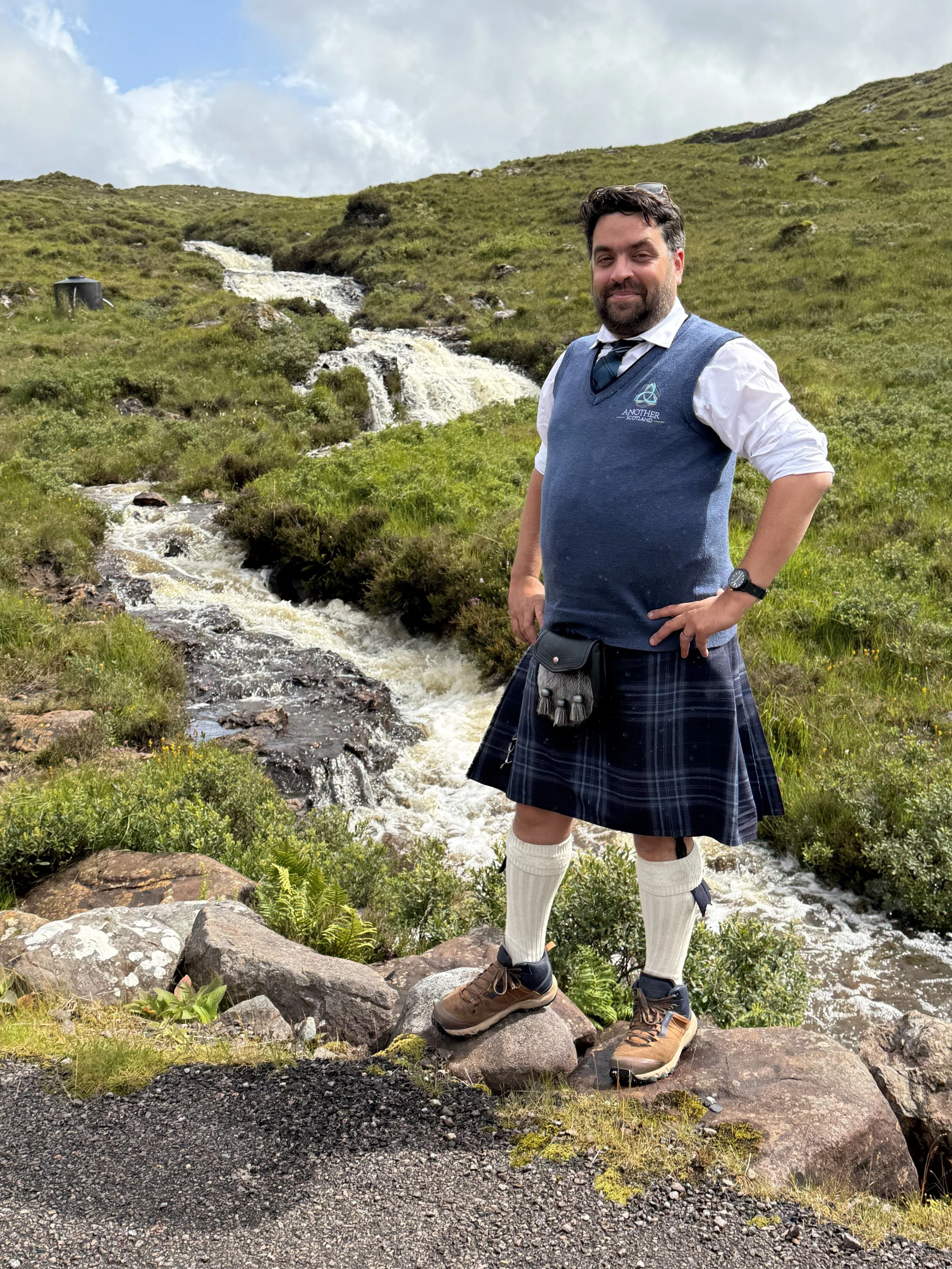 Man in traditional Scottish attire standing on rocks beside a stream in a green mountainous area.