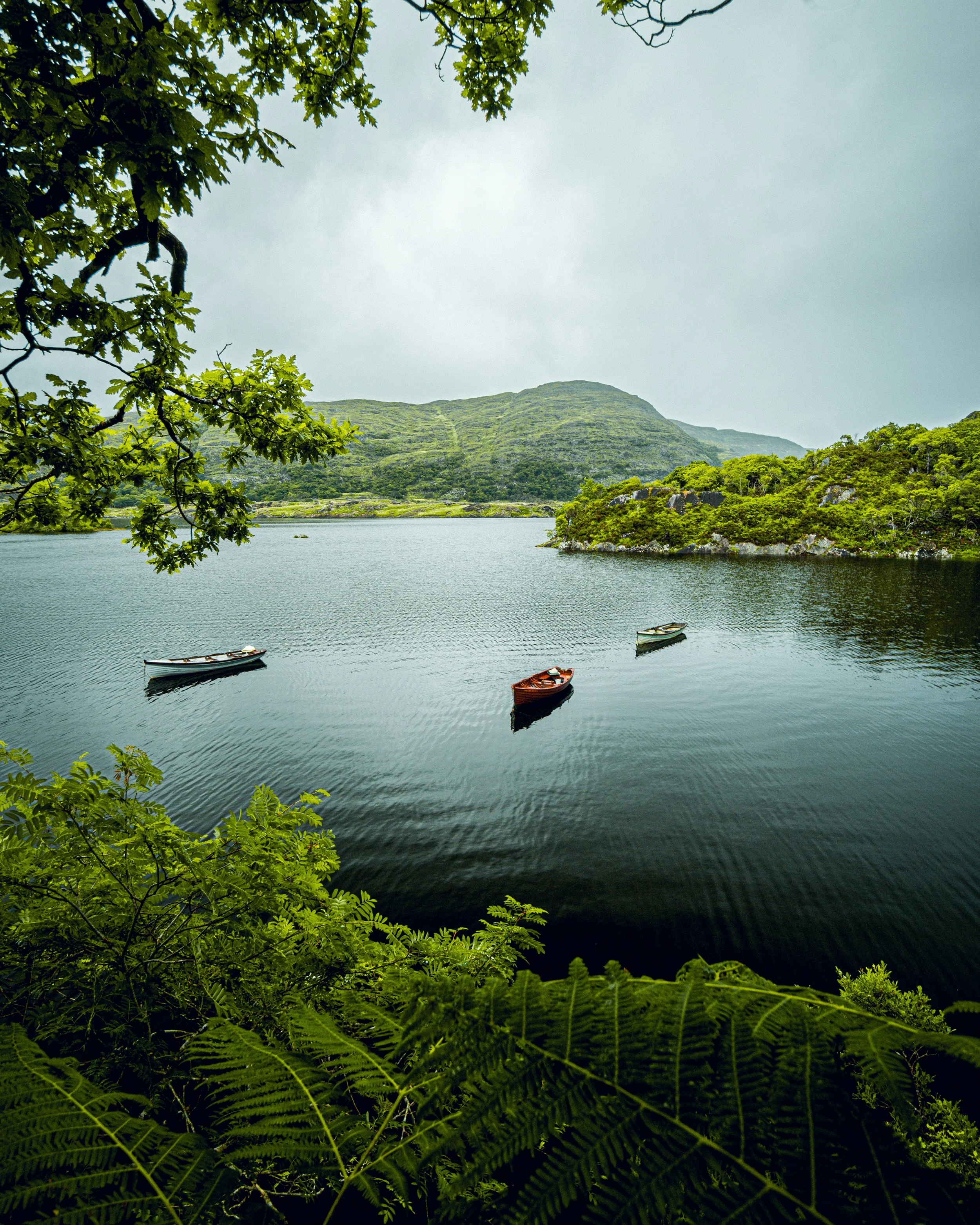 A peaceful lake surrounded by lush green trees and hills, with three small boats floating on the water under a cloudy sky.