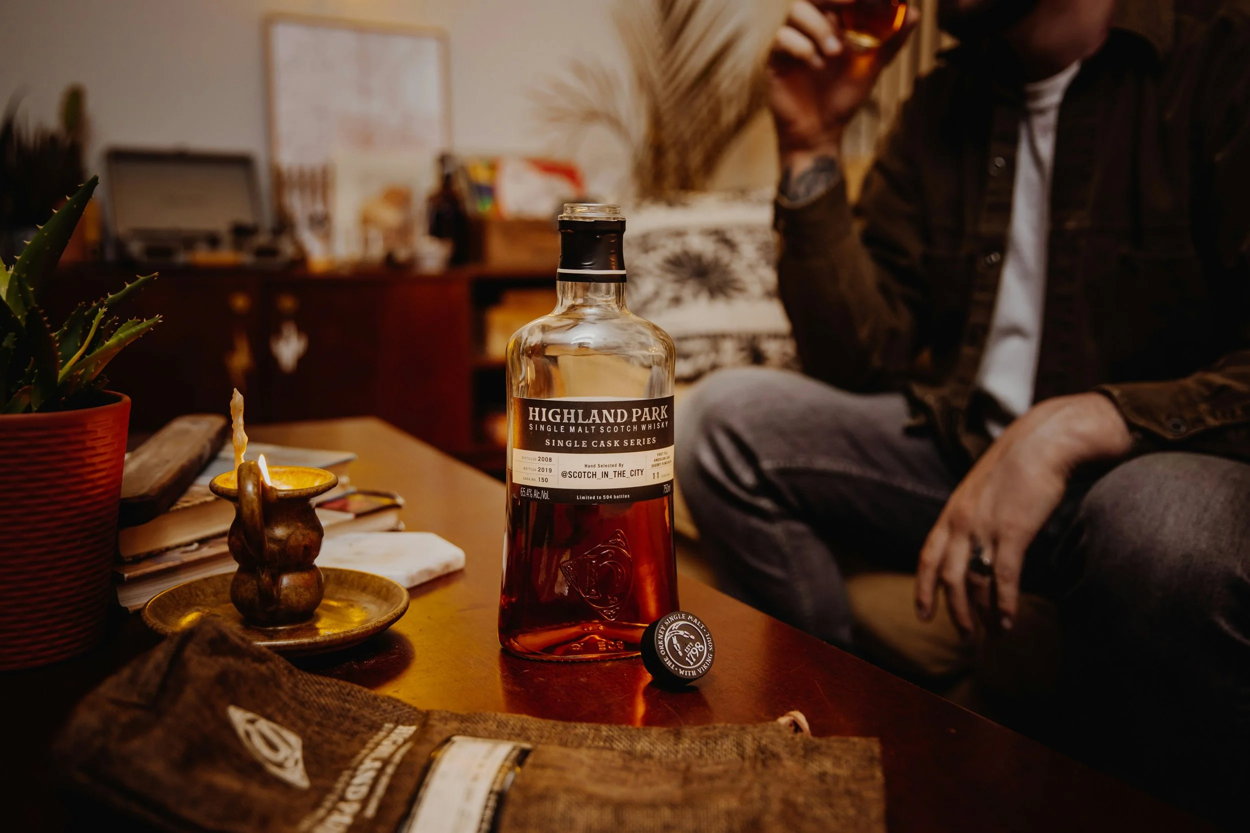 Bottle of Highland Park single malt Scotch whisky on a wooden table next to a lit candle, potted plant, and a small plate. In the background, two people are sitting and drinking, with one person wearing a brown jacket and the other with blonde hair and patterned sweater.
