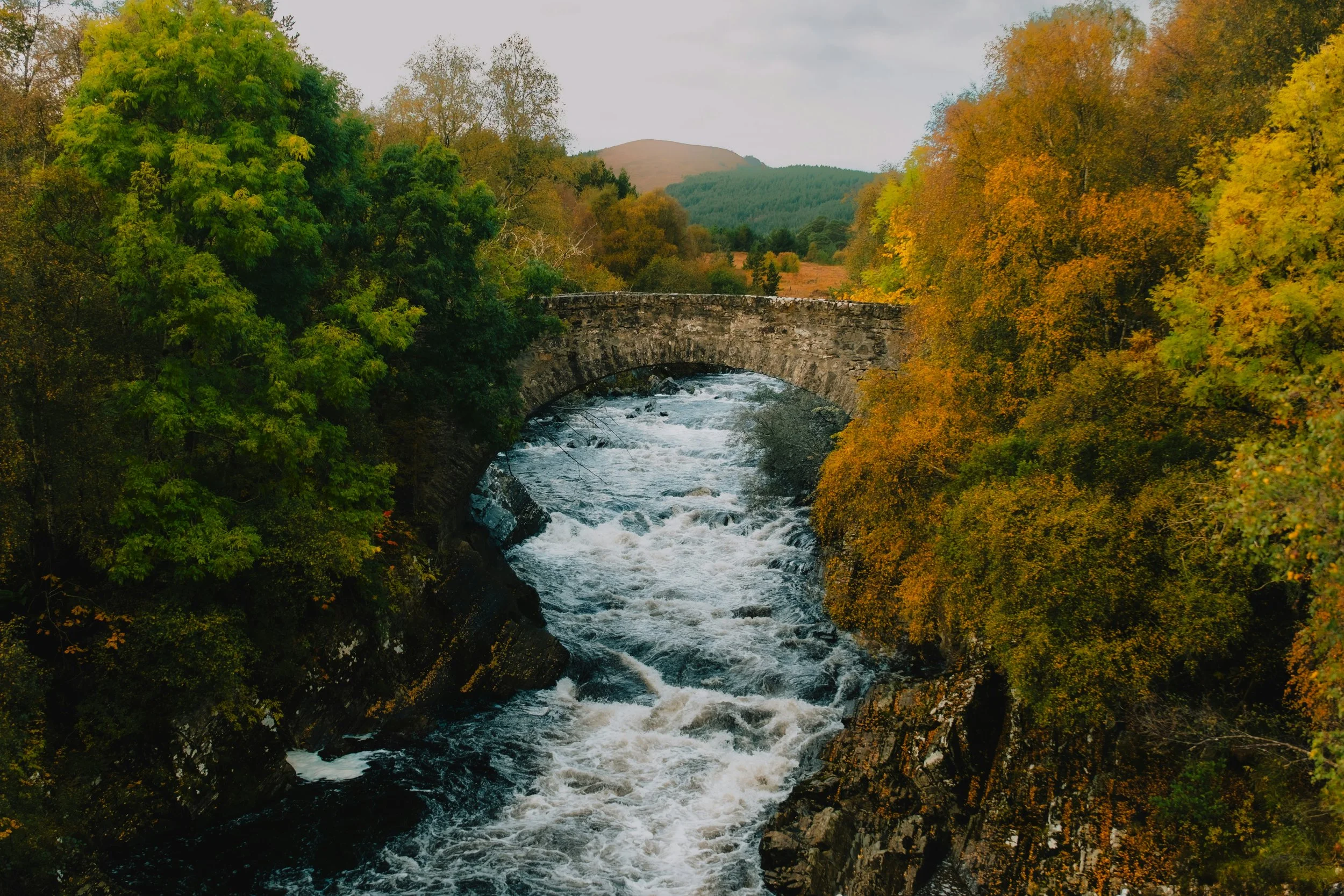 A stone bridge over a rushing river surrounded by trees with green and orange fall foliage.