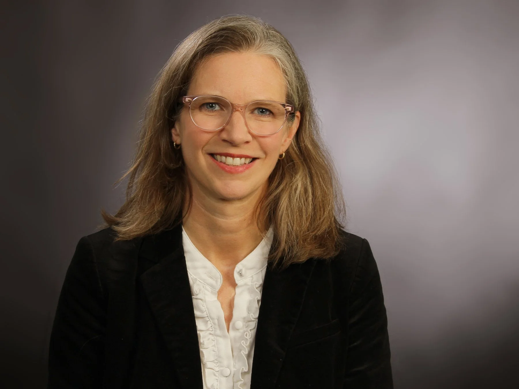 A woman with shoulder-length blonde hair, wearing glasses, a white blouse, and a black blazer, smiling against a gray gradient background.
