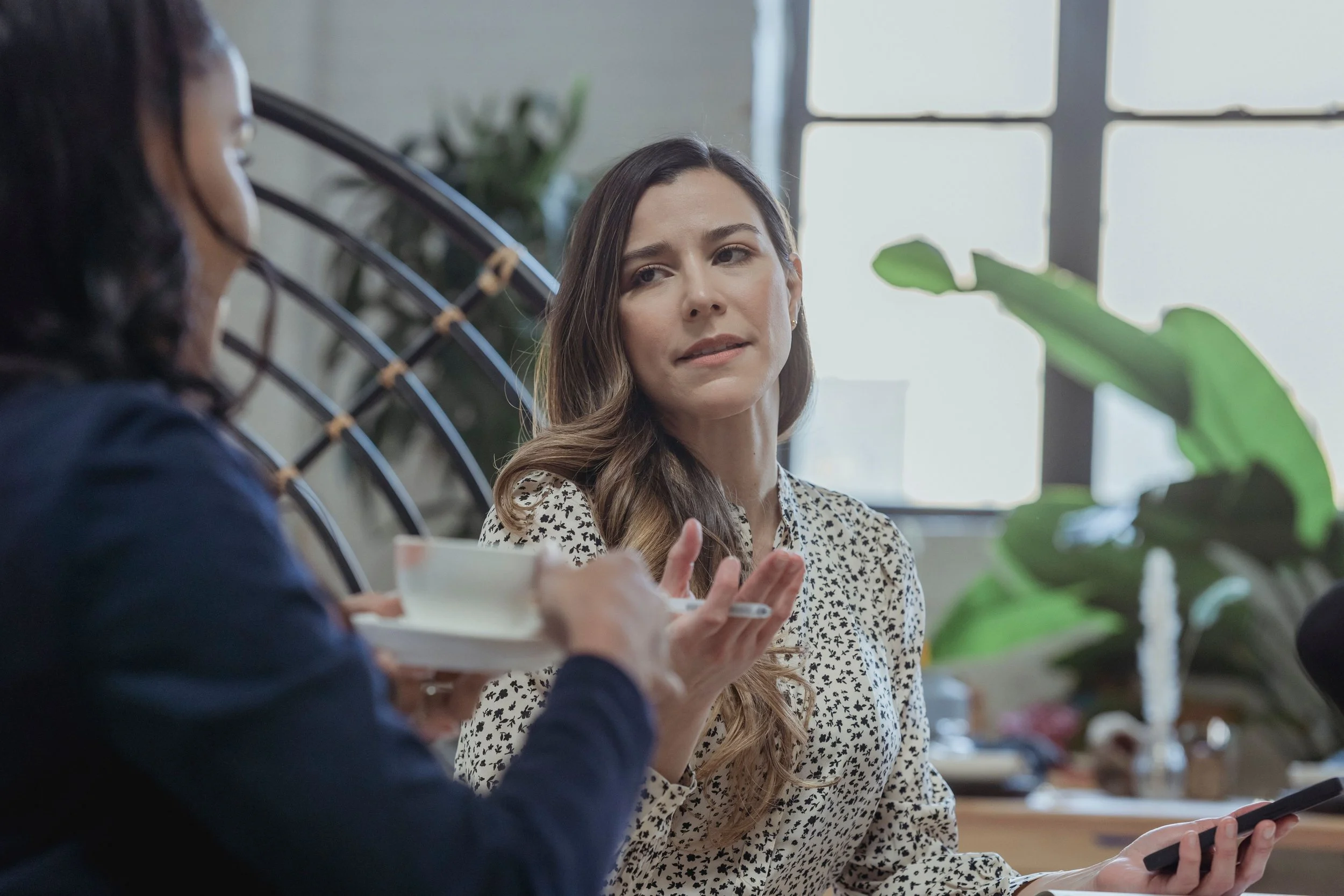 Two women sitting at a cafe table, with one looking slightly disappointed while the other looks down thoughtfully, practicing the uncomfortable feeling of not immediately fixing someone else's emotions.