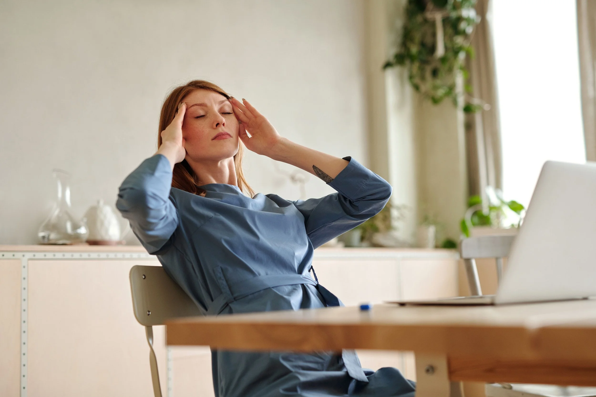 A capable, put-together woman standing in her kitchen, looking overwhelmed and quietly exhausted as she rubs her temple while reviewing a busy family calendar and several to-do lists.
