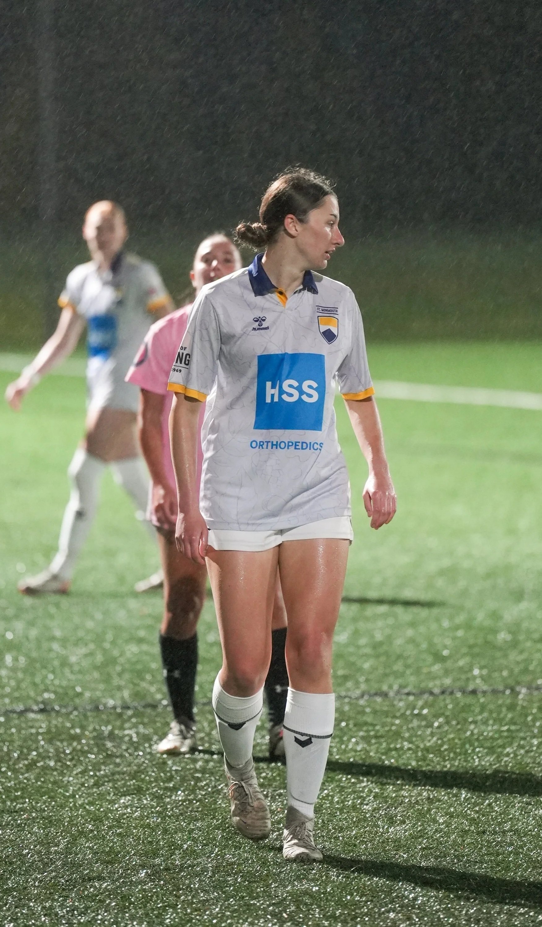 A female soccer player in a gray jersey with blue and yellow accents stands on a wet field at night, with her hair tied back, followed by other players partially visible behind her.