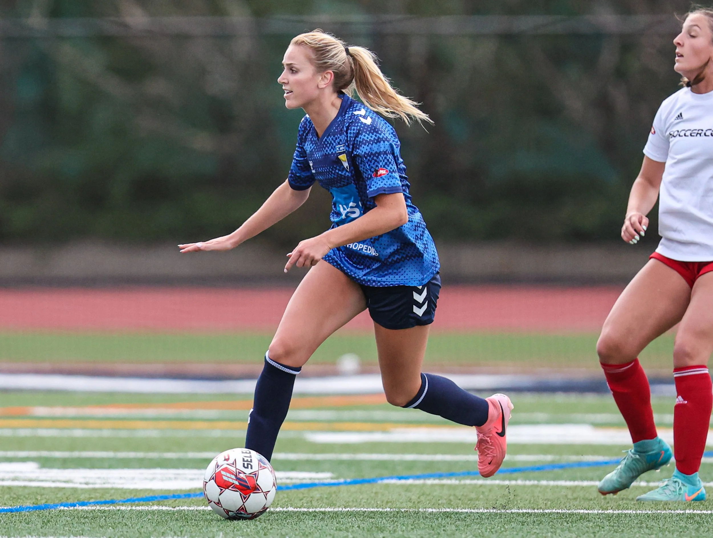 A female soccer player in a blue jersey and black shorts is running on a sports field, about to kick a soccer ball. Another female player in a white shirt and red shorts is visible nearby.