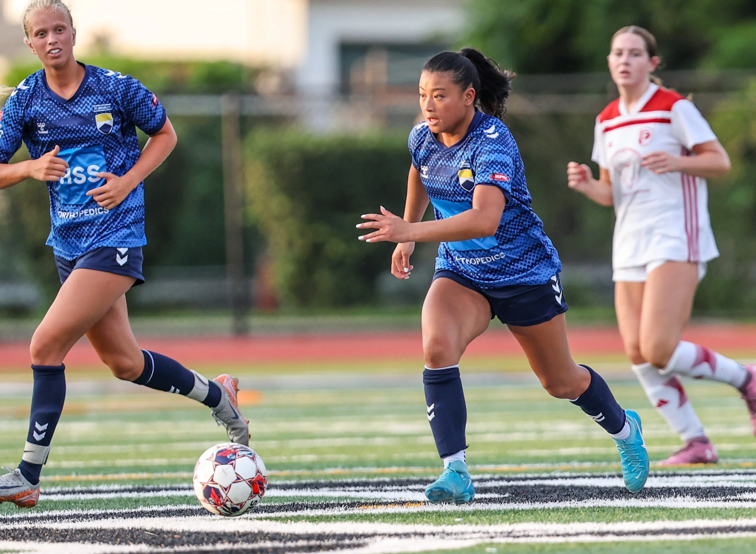 Girls playing soccer on a field, with a girl in a blue jersey controlling the ball, and other players nearby.