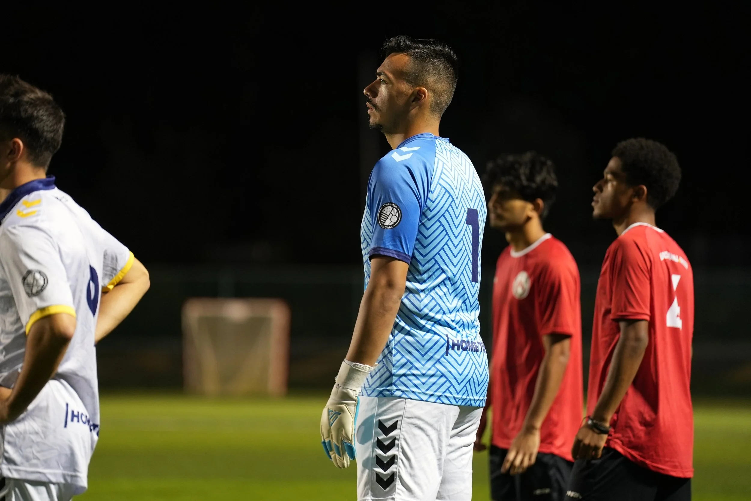 Soccer players standing on a field at night, wearing team jerseys, with one in a blue goalkeeper uniform and others in red jerseys.