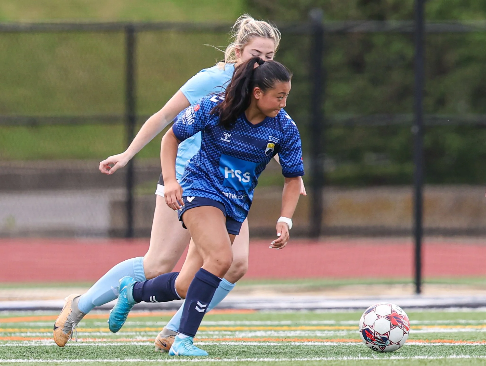 Female soccer player kicking a yellow soccer ball on the field during practice or game, with another player and team bench in the background.
