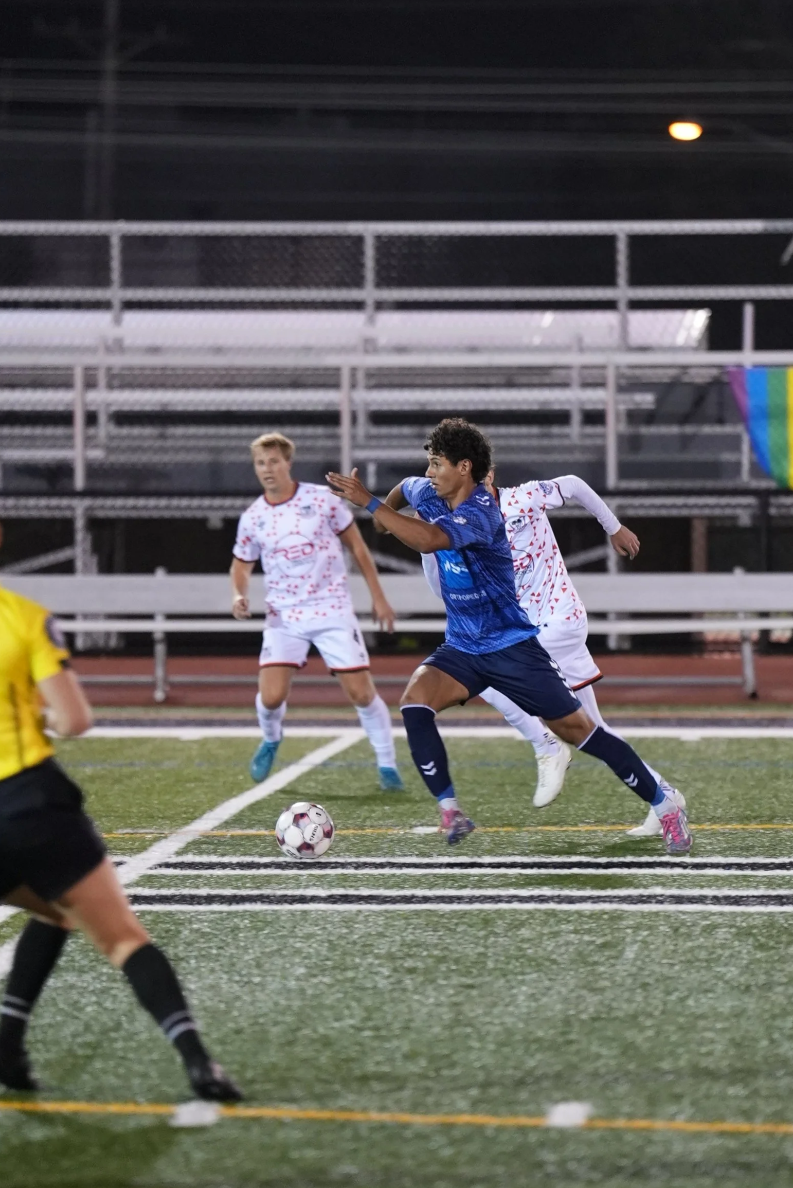 A soccer match with players on the field, focusing on a player in a blue uniform running towards the ball while two players in white uniforms approach from behind, with a referee in a yellow shirt nearby.