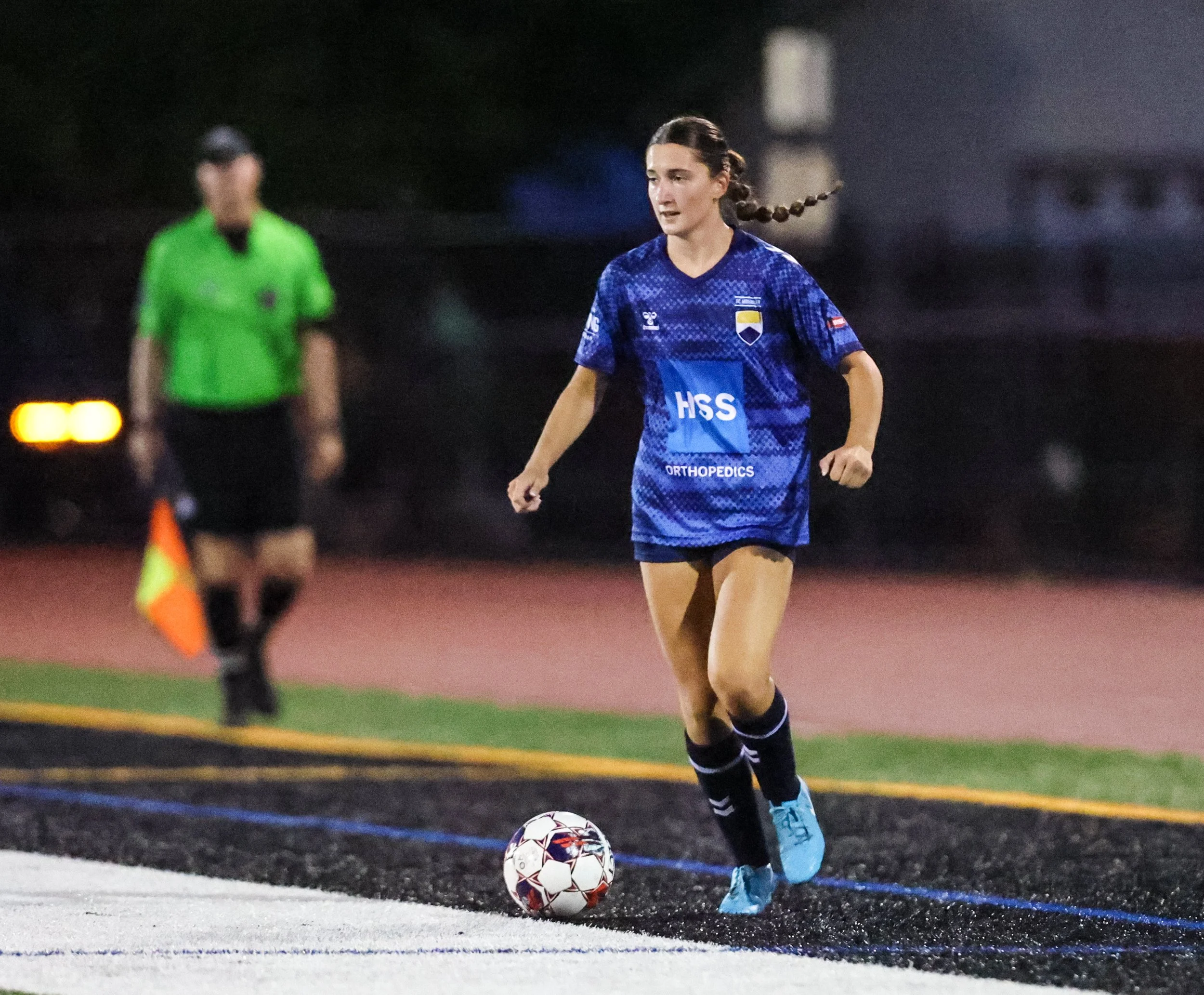 A young female soccer player in a blue jersey dribbling a soccer ball on a field at night, with a referee in a green shirt in the background.