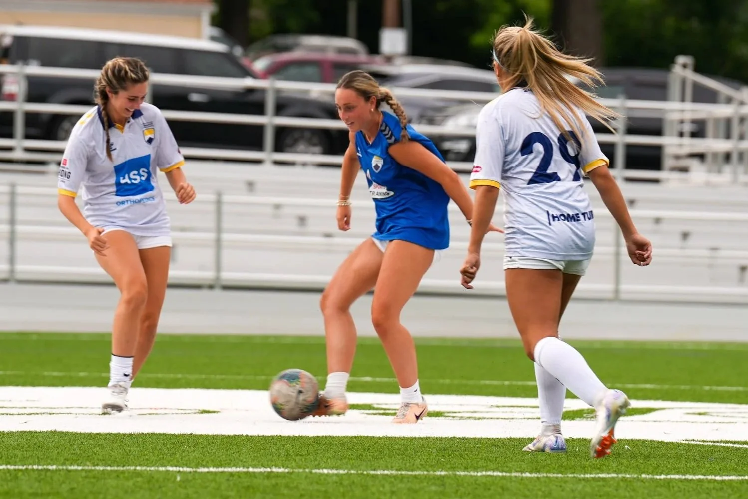 Three women playing soccer on a field, two in white jerseys and one in blue, with a soccer ball near them.