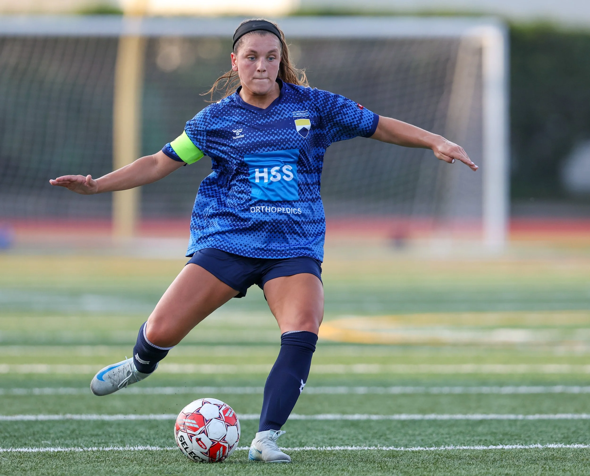 A female soccer player wearing a blue jersey with a captain's armband, black shorts, and socks, kicking a soccer ball on a field with a goal in the background.