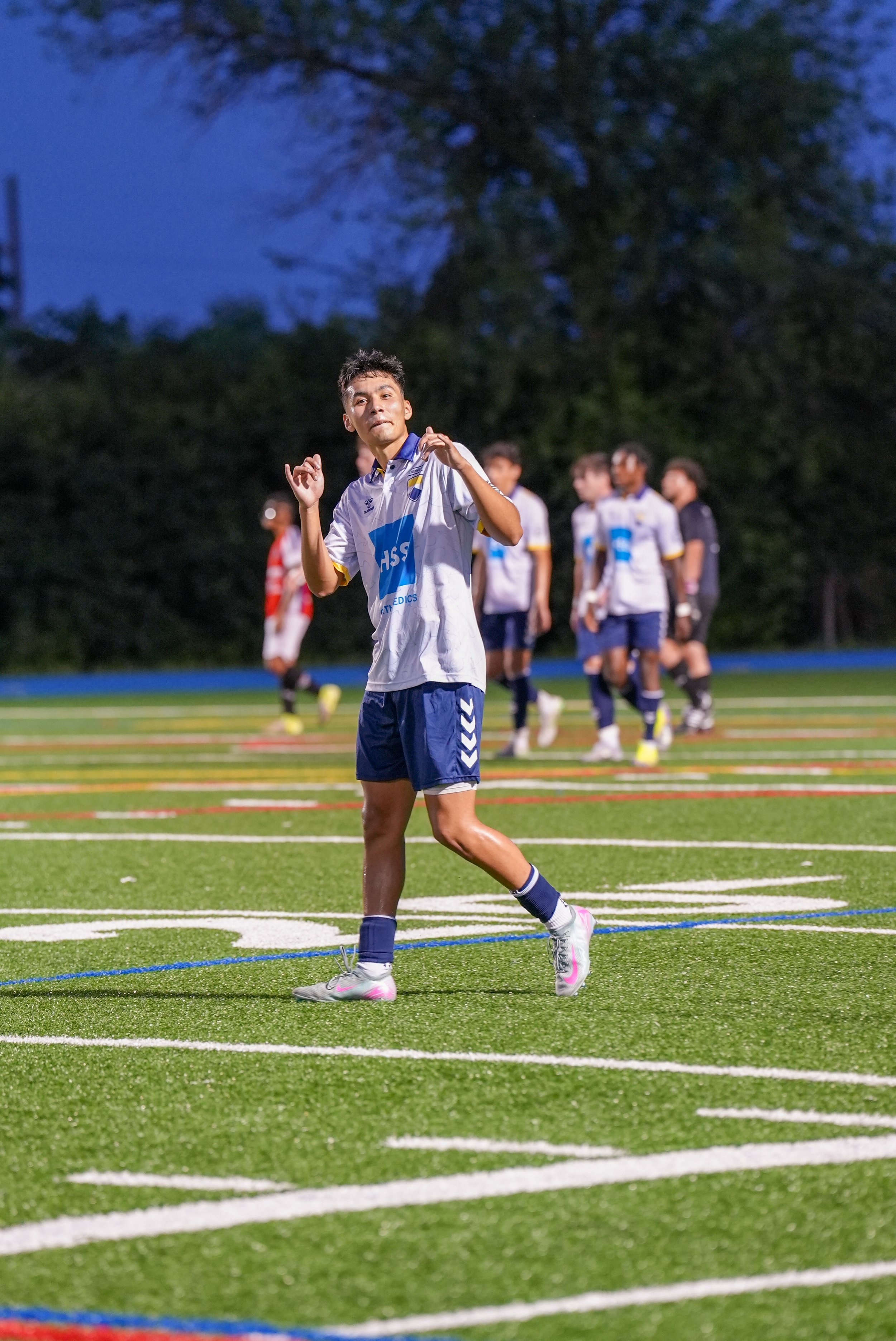 A soccer player in a white and blue uniform on the field at night with other players in the background.