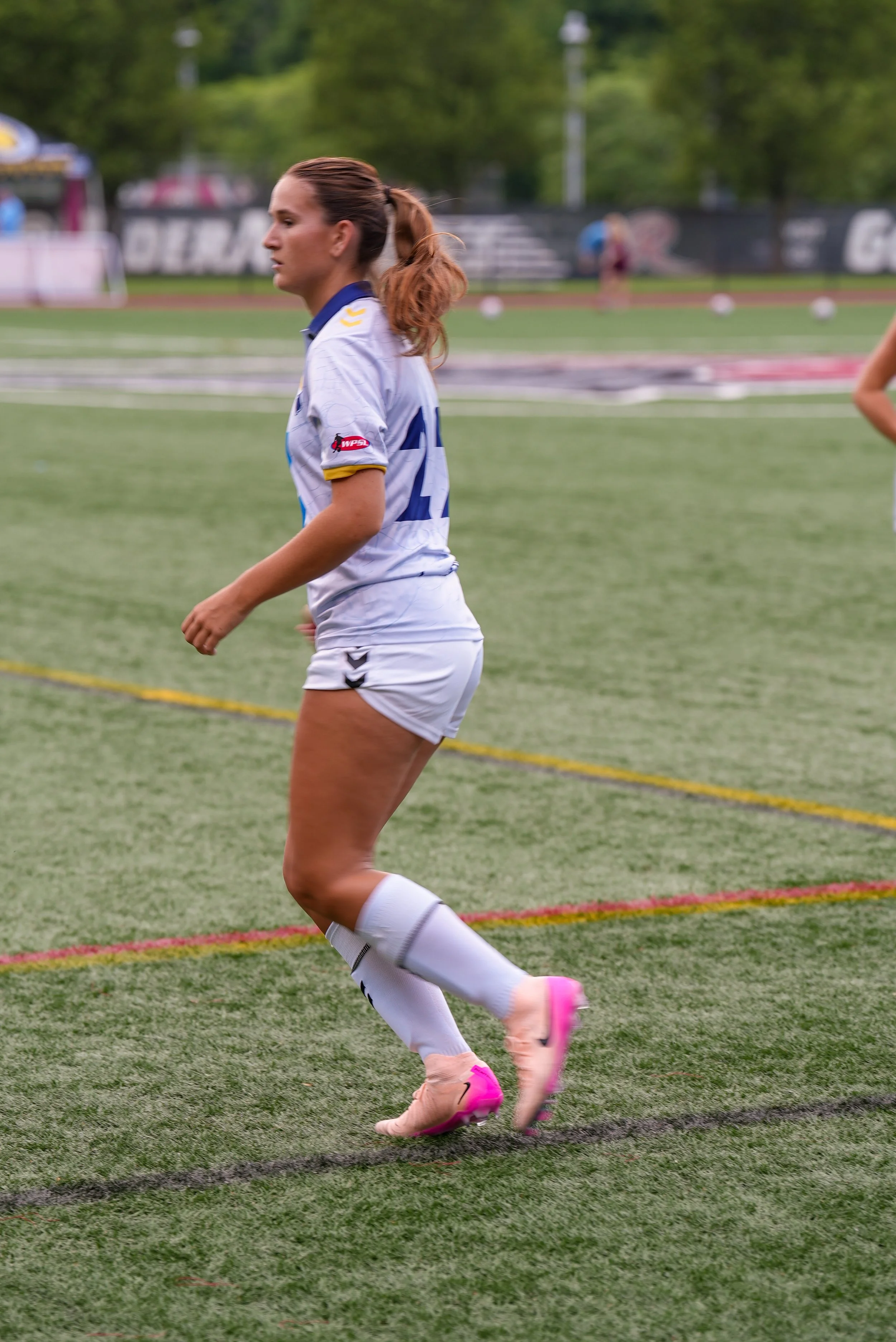 A female soccer player in a white and blue uniform, pink cleats, and long soccer socks standing on a green soccer field during a game.