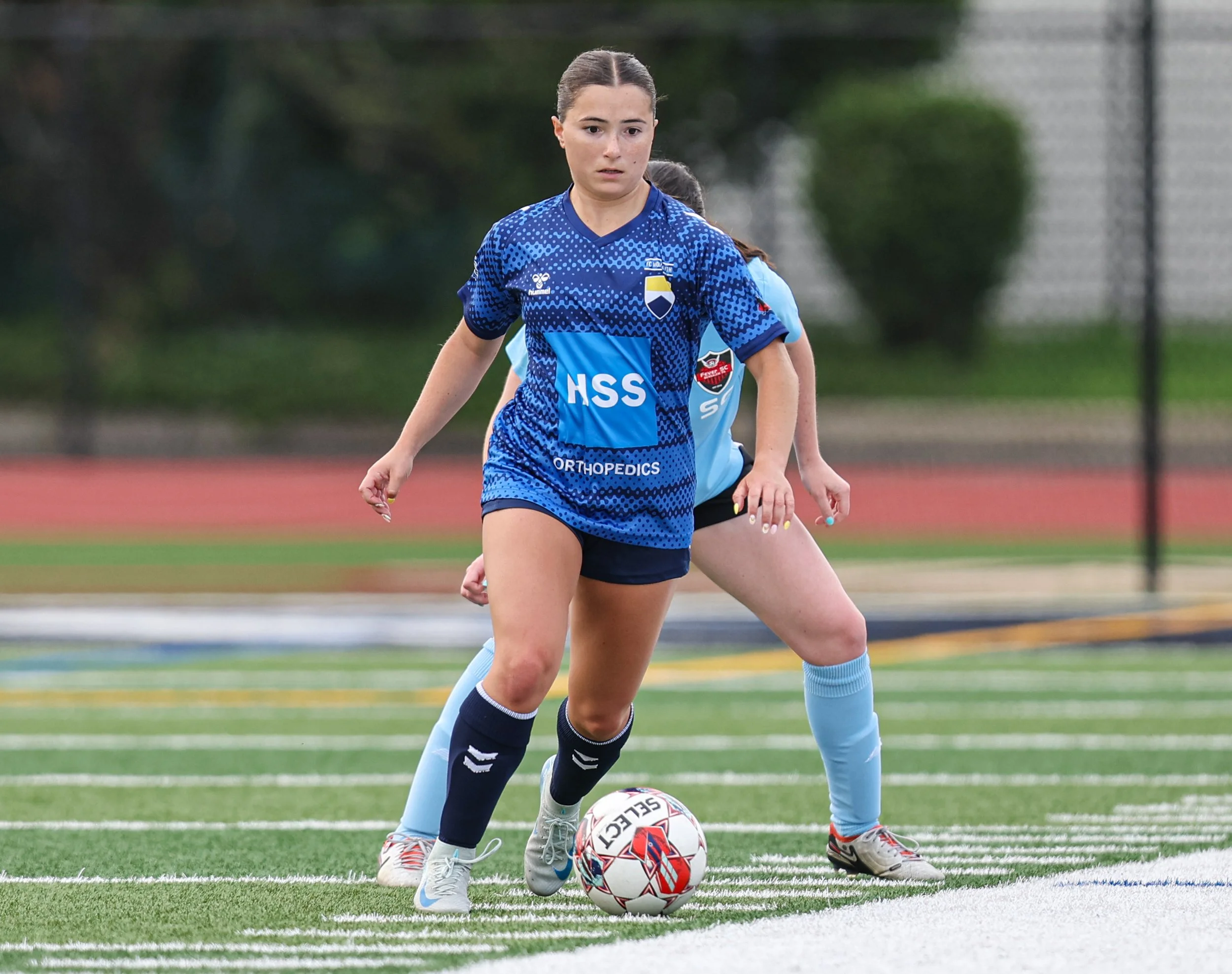 A female soccer player in a blue and black uniform controlling a soccer ball during a game on a green field.