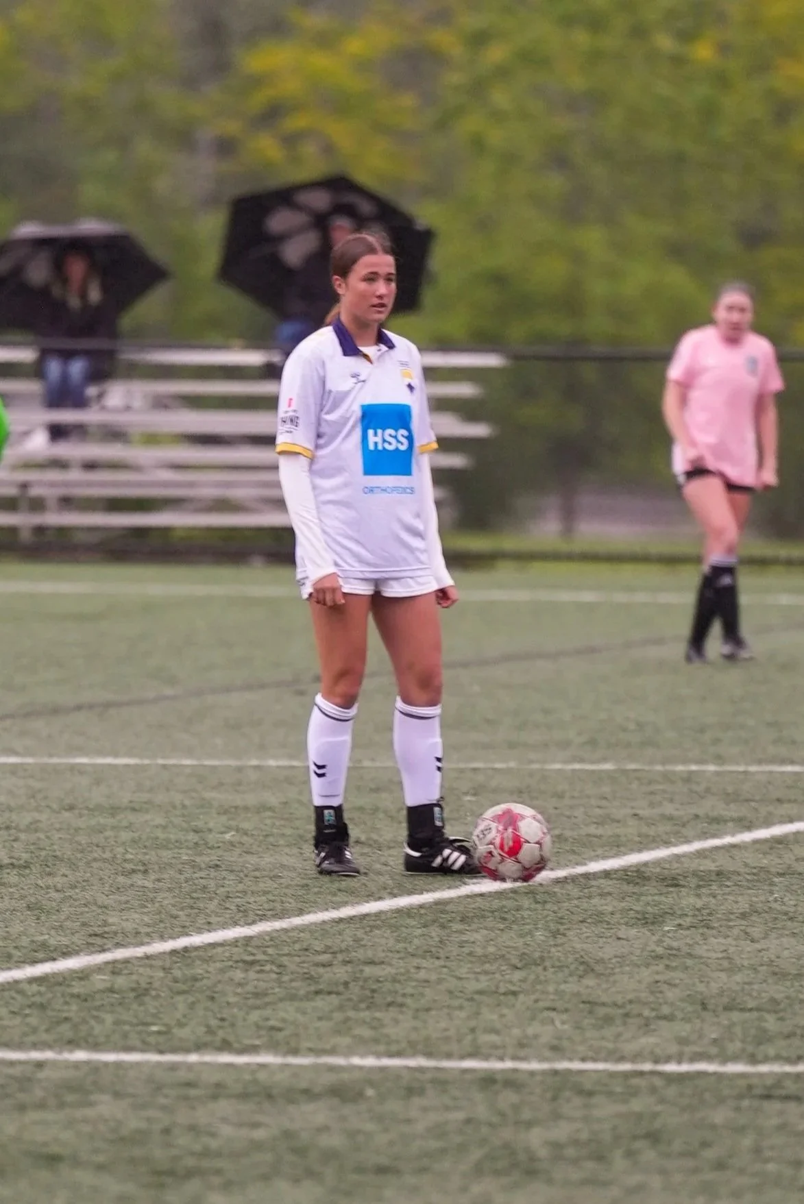 A young woman in a soccer uniform standing on a field with a soccer ball at her feet, while other people with umbrellas are in the background.