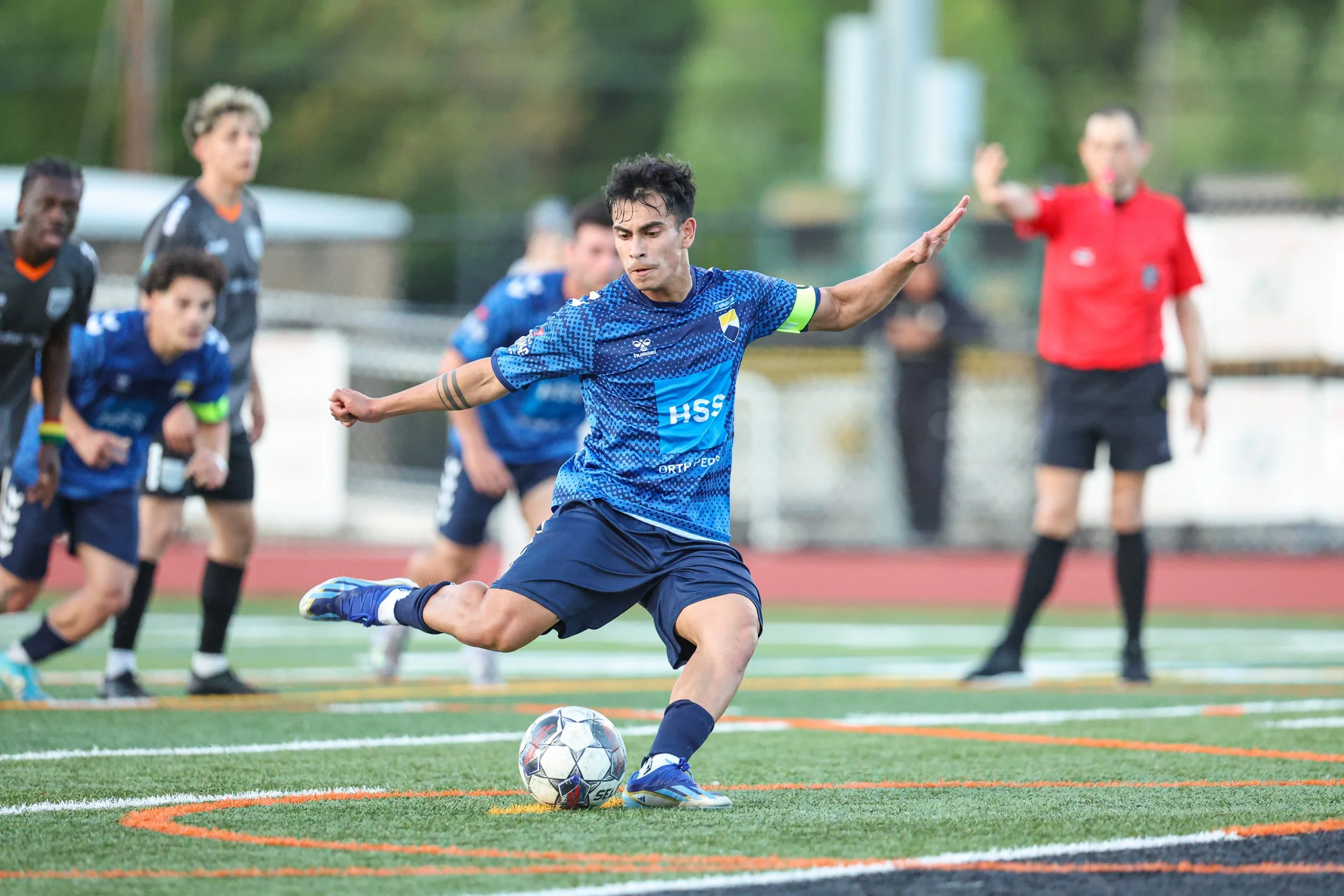A soccer player in a blue jersey kicks a soccer ball during a match on a field, with other players, a referee in red, and a fence in the background.