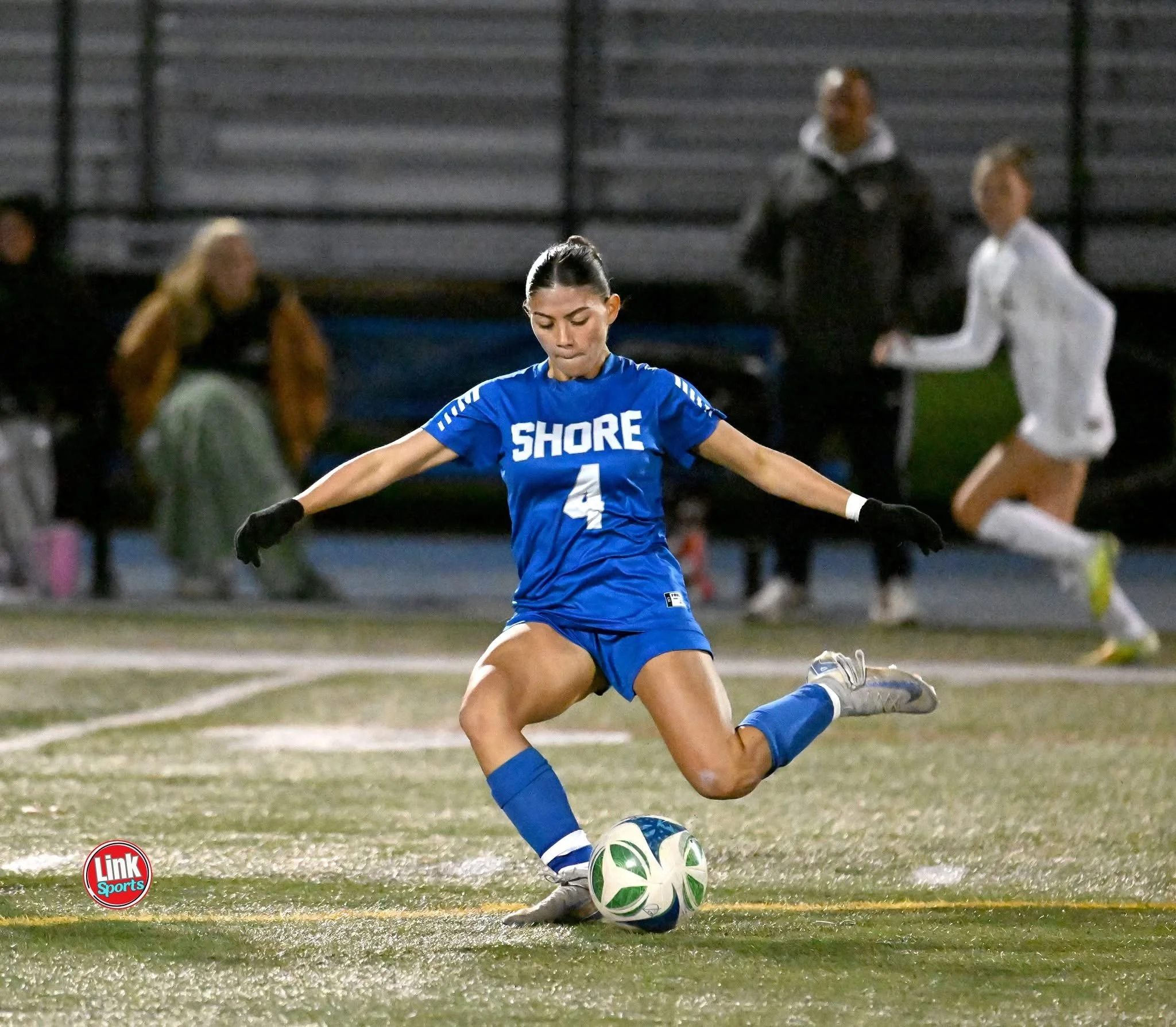 A female soccer player in a gray jersey with blue and yellow accents stands on a wet field at night, with her hair tied back, followed by other players partially visible behind her.
