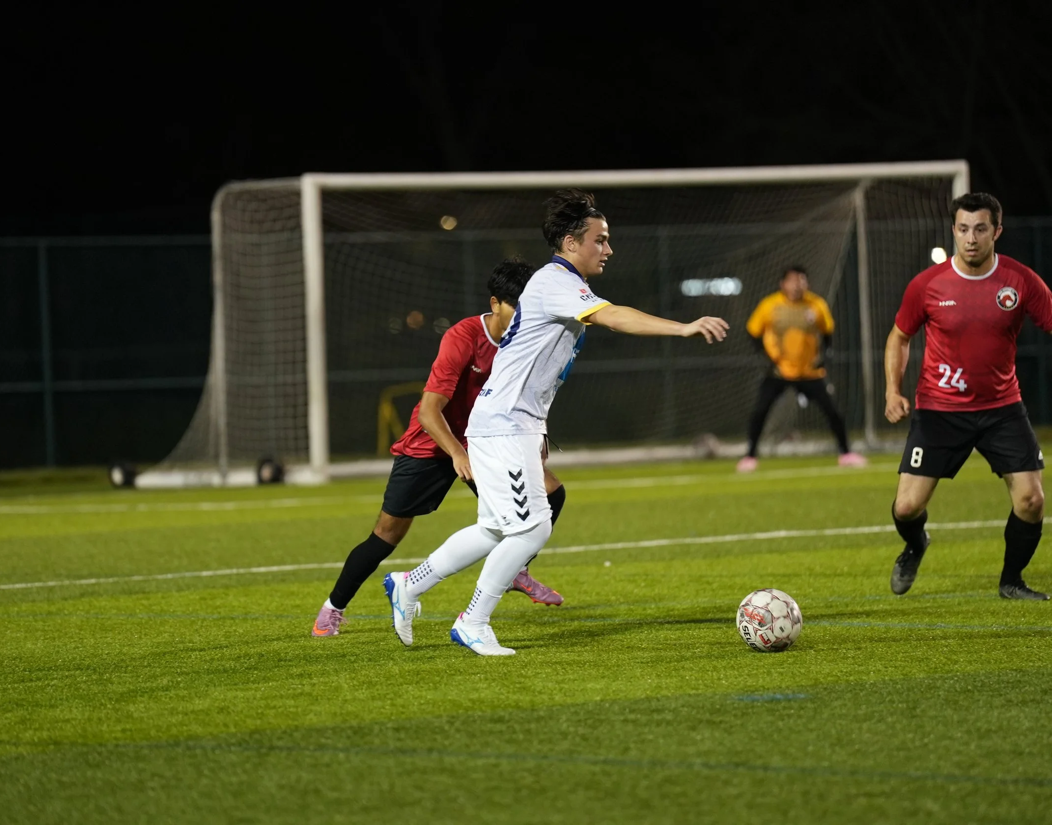 Soccer players contest for the ball on a field at night, with a goalkeeper standing in front of the goal in the background.