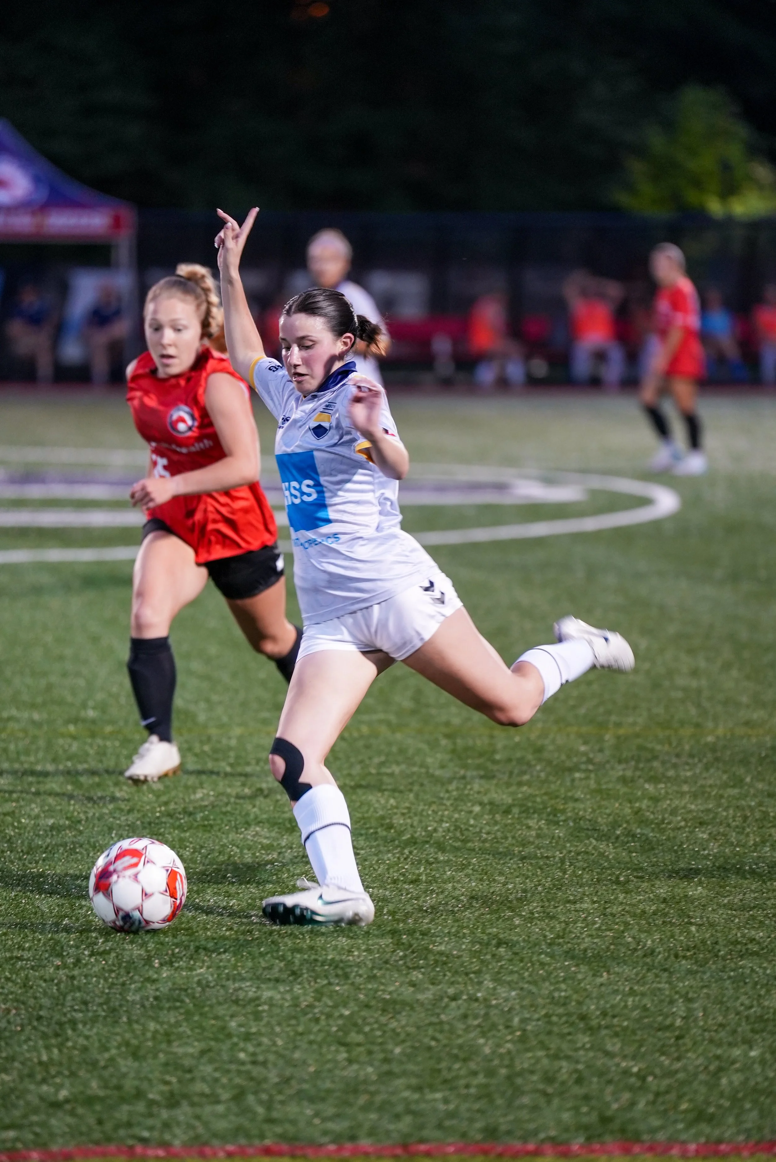A women’s soccer game with a player in a white uniform kicking a ball while an opponent in a red uniform runs nearby on a green field during dusk.