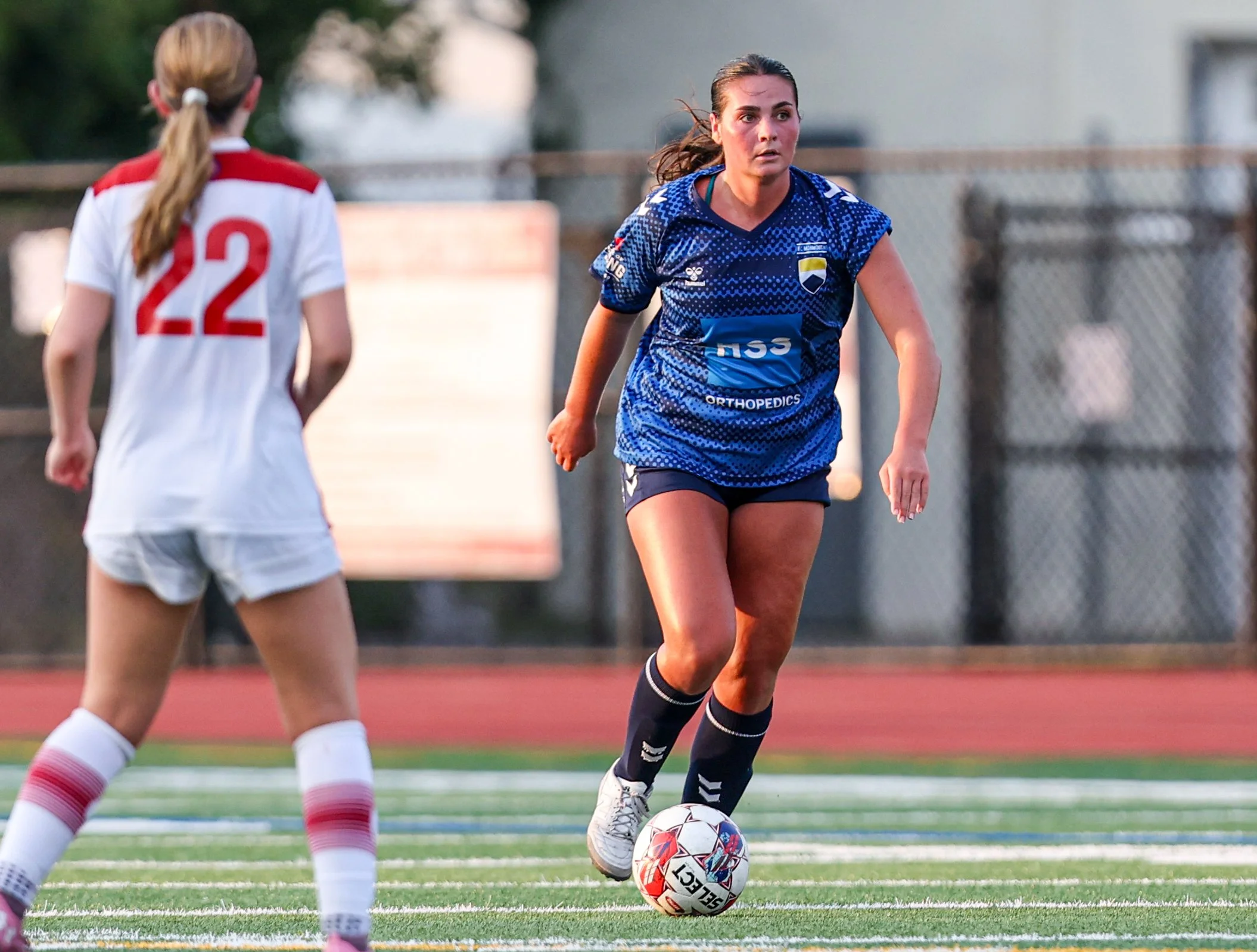 A female soccer player in a blue jersey dribbling a soccer ball on a field while an opponent in a white and red jersey watches.