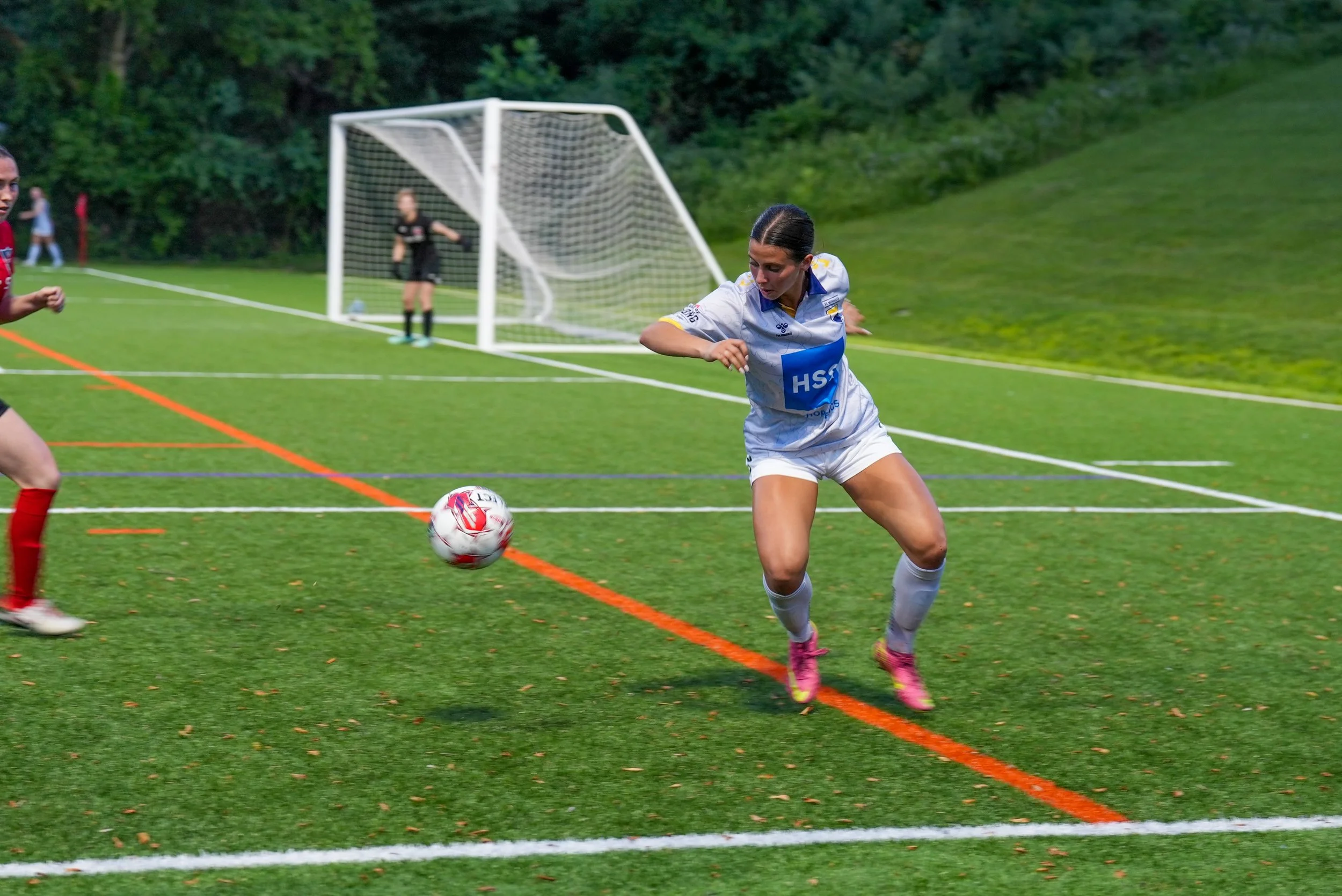 Female soccer player in white uniform with pink cleats chasing a soccer ball on a green field during a game, with a goalkeeper in black in front of the goal in the background.