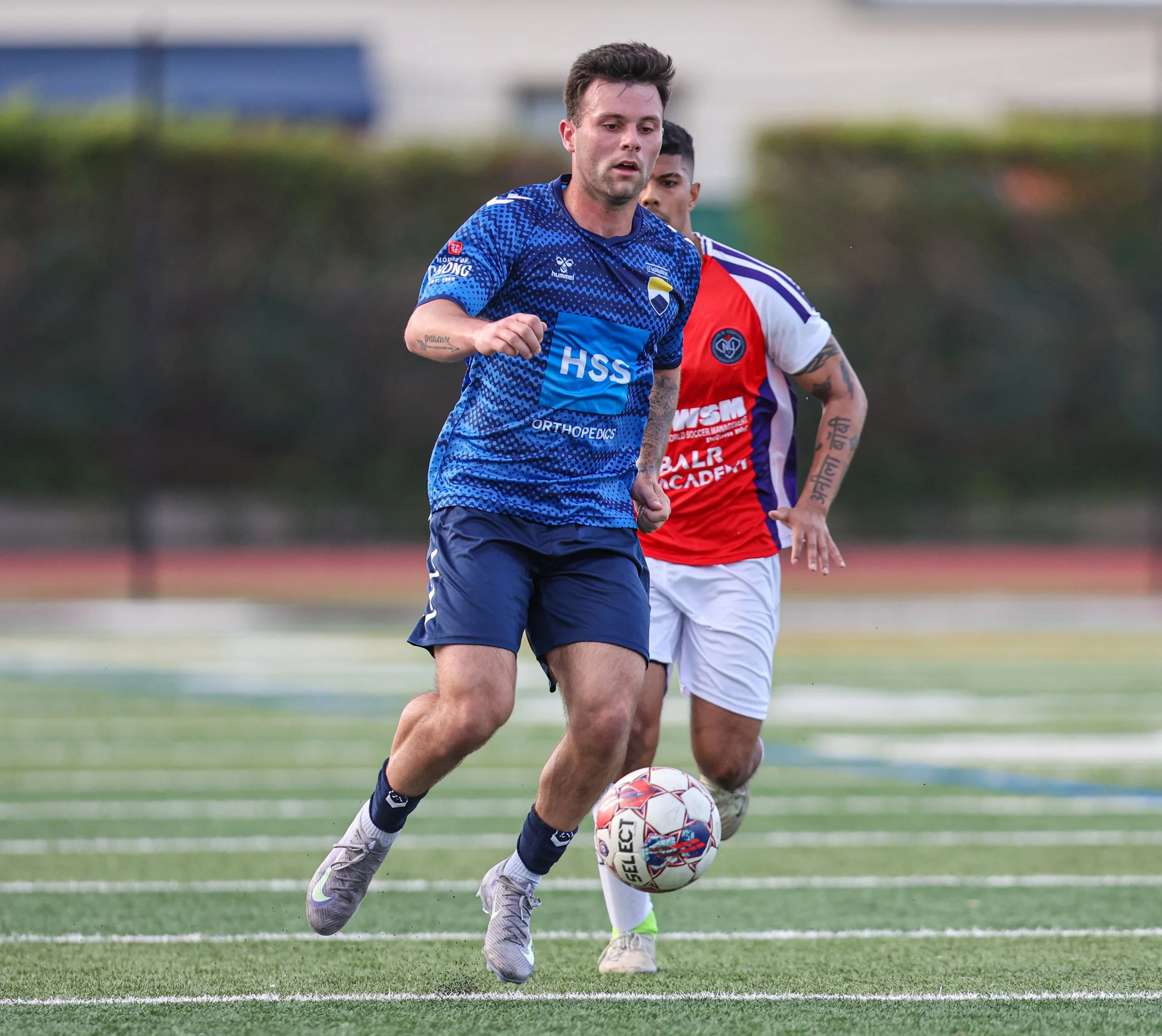 Two soccer players running on the field, one in a blue jersey and the other in a red and white jersey, with a soccer ball.