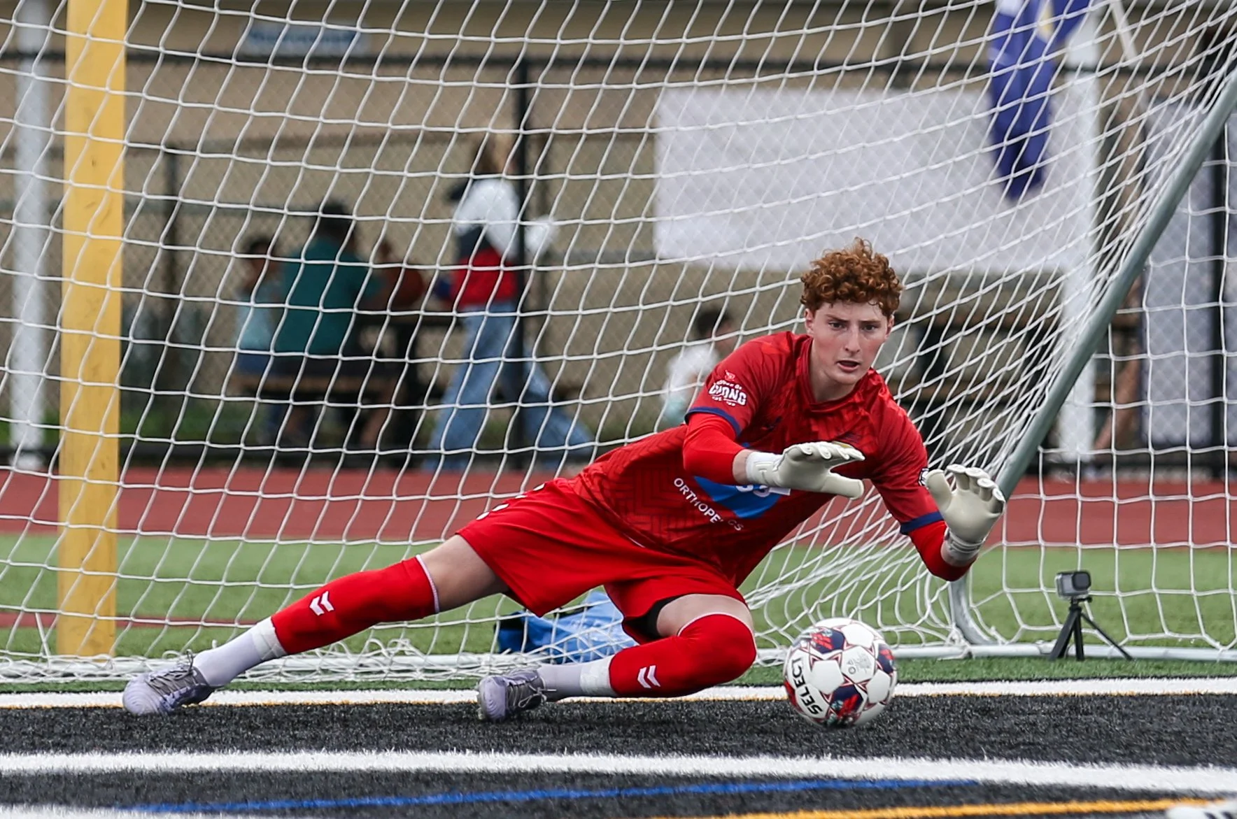 A young male soccer goalkeeper in red uniform diving to make a save during a match, with a soccer ball near his hands inside the goal.