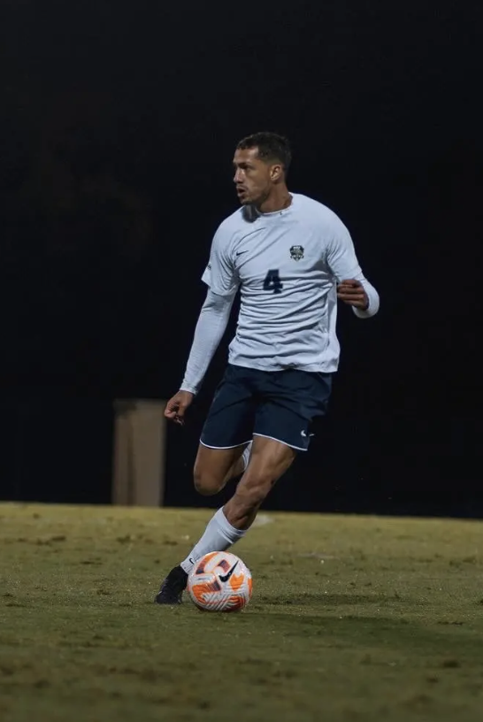A soccer match with players on the field, focusing on a player in a blue uniform running towards the ball while two players in white uniforms approach from behind, with a referee in a yellow shirt nearby.