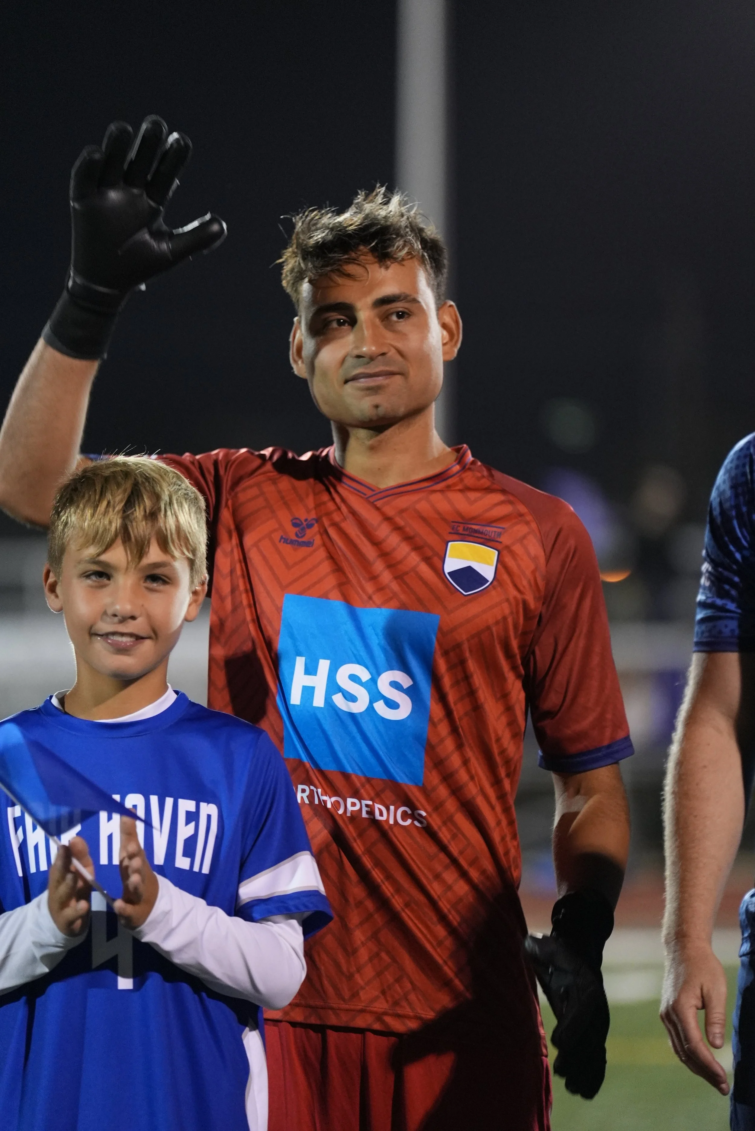 A young boy and a soccer player waving at the camera during a soccer match at night. The boy is wearing a blue soccer jersey. The player is in a red uniform, wearing black gloves, and has tousled hair.