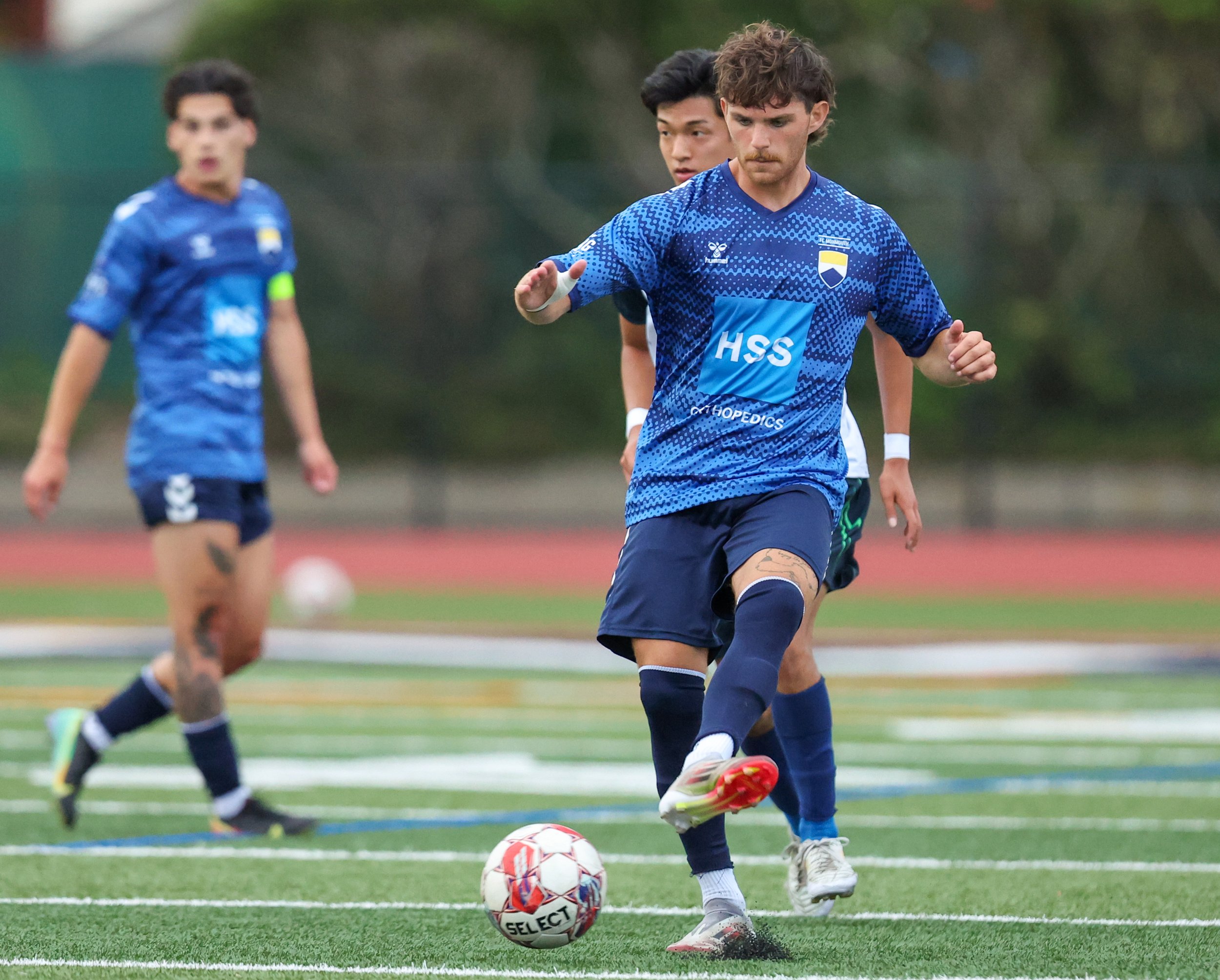 A group of young men playing soccer on a field during daytime, with one player preparing to kick the ball while others are in the background.