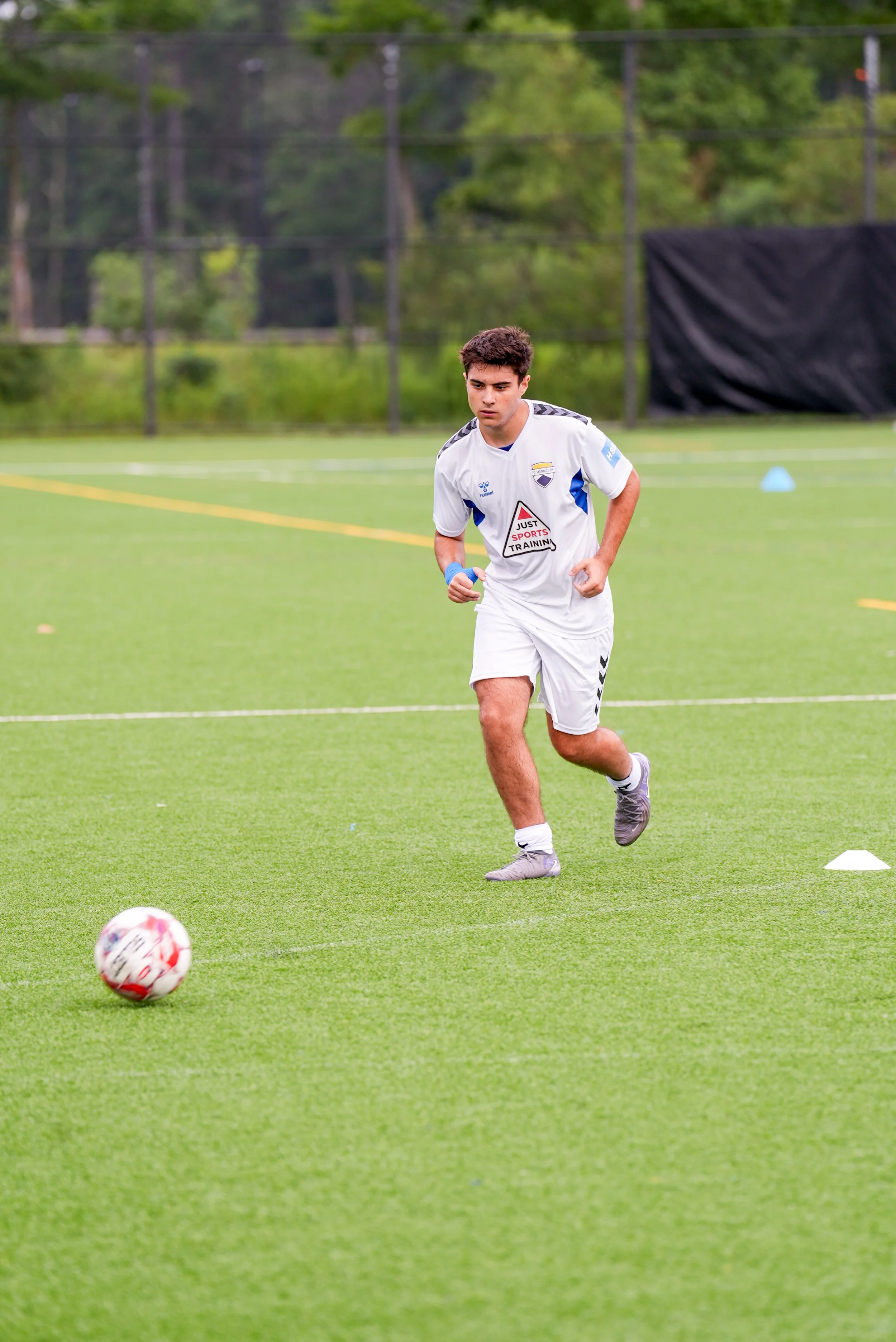 Young male soccer player in white uniform running on field with soccer ball nearby during practice.