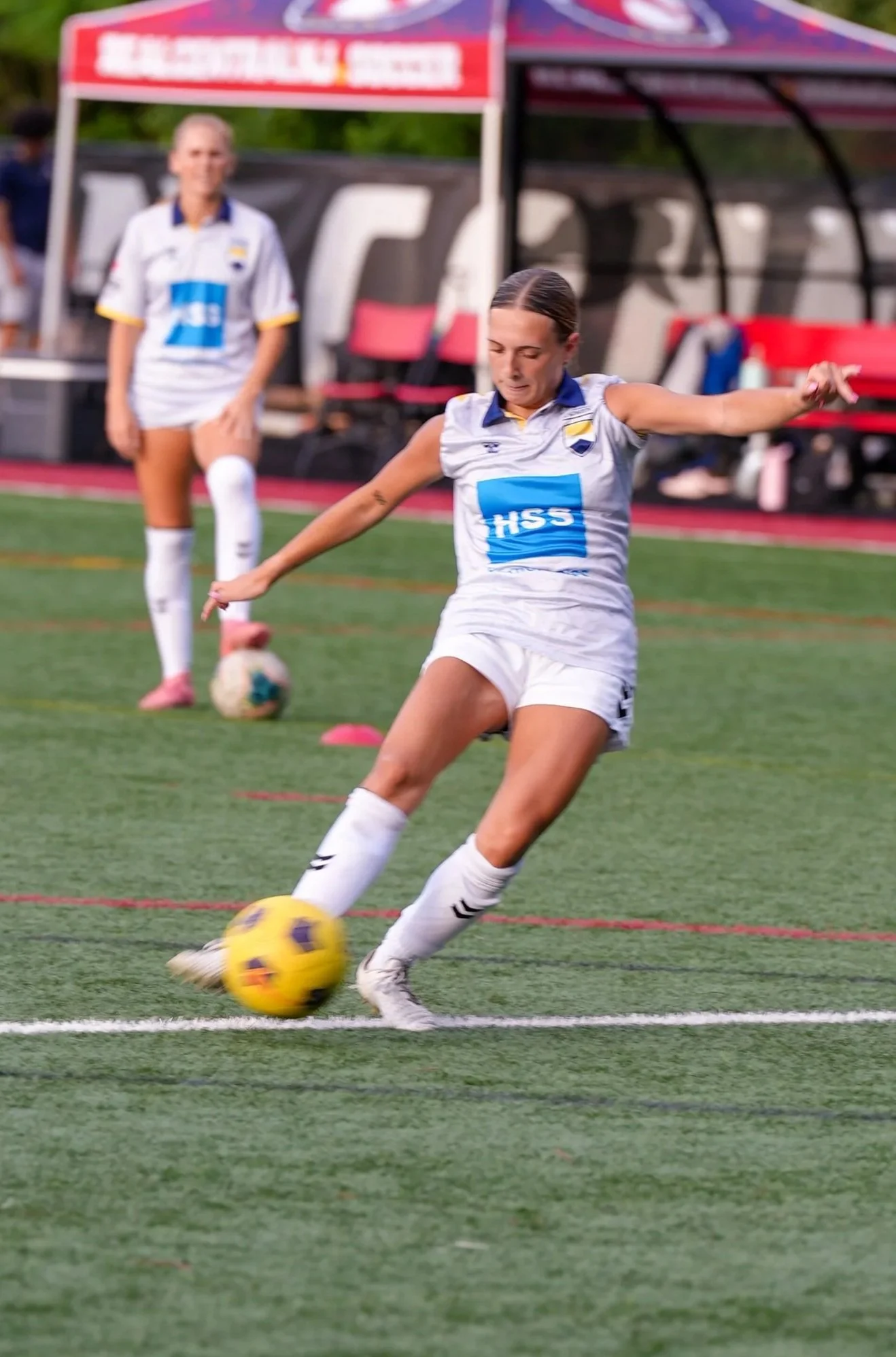 Female soccer player kicking a yellow soccer ball on the field during practice or game, with another player and team bench in the background.