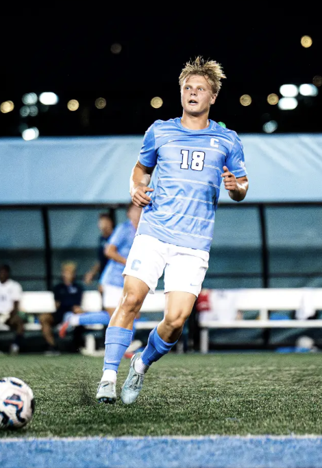 A soccer player in a blue jersey kicks a soccer ball during a match on a field, with other players, a referee in red, and a fence in the background.