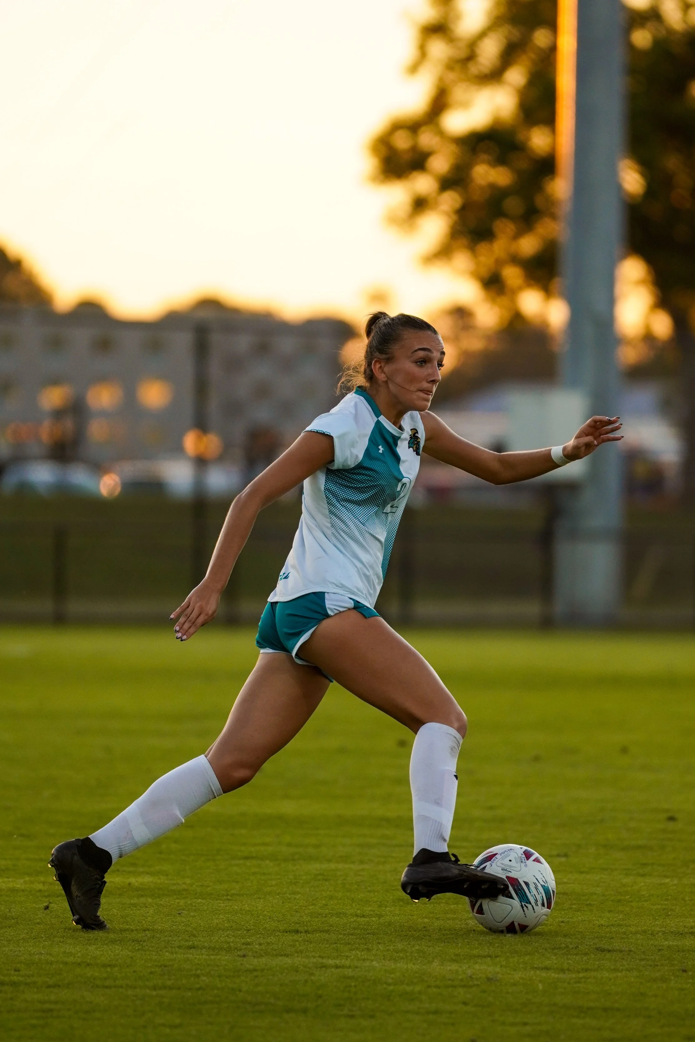 A female soccer player wearing a blue jersey with a captain's armband, black shorts, and socks, kicking a soccer ball on a field with a goal in the background.