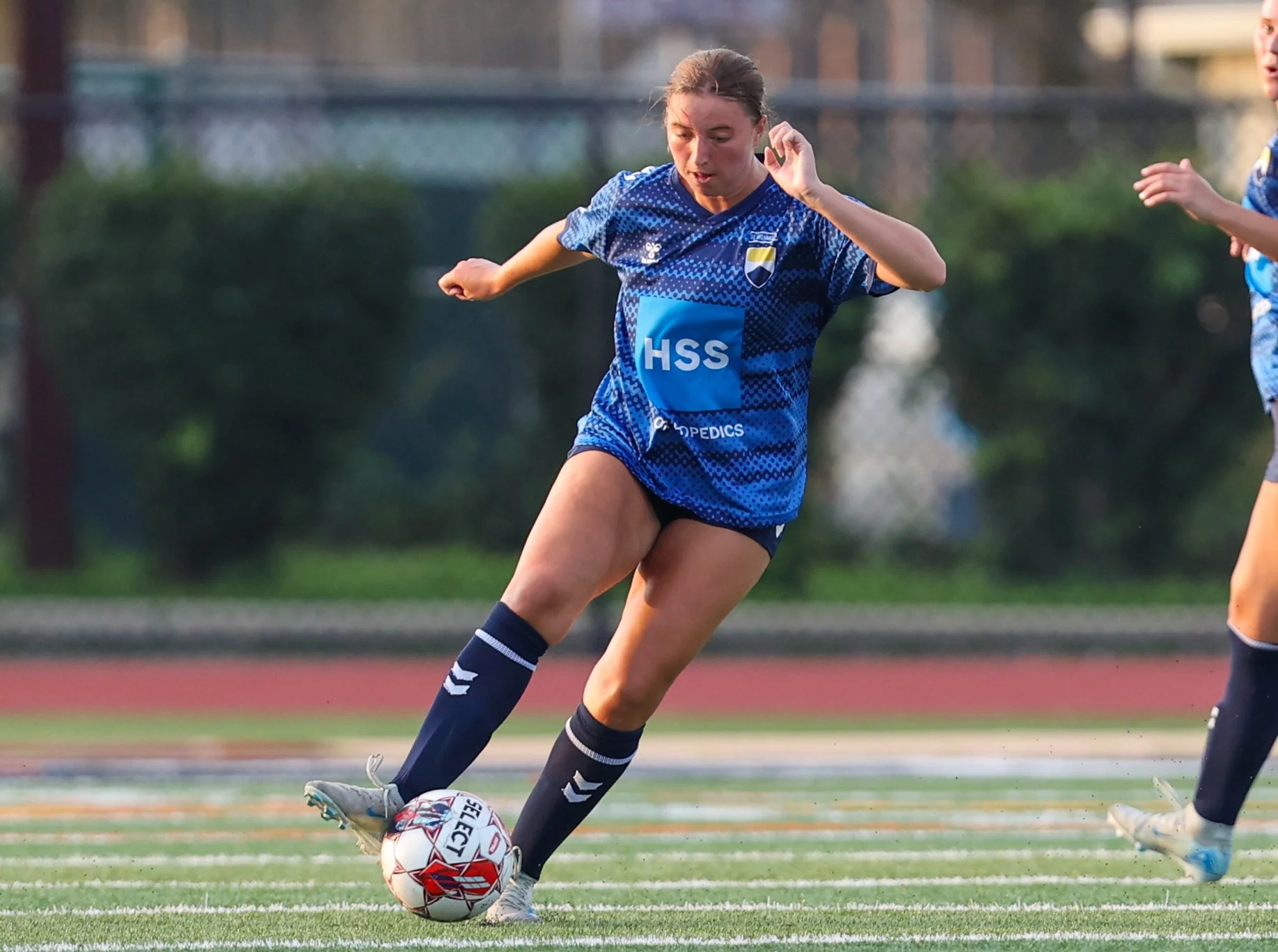 A female soccer player in blue uniform kicking a soccer ball on the field.